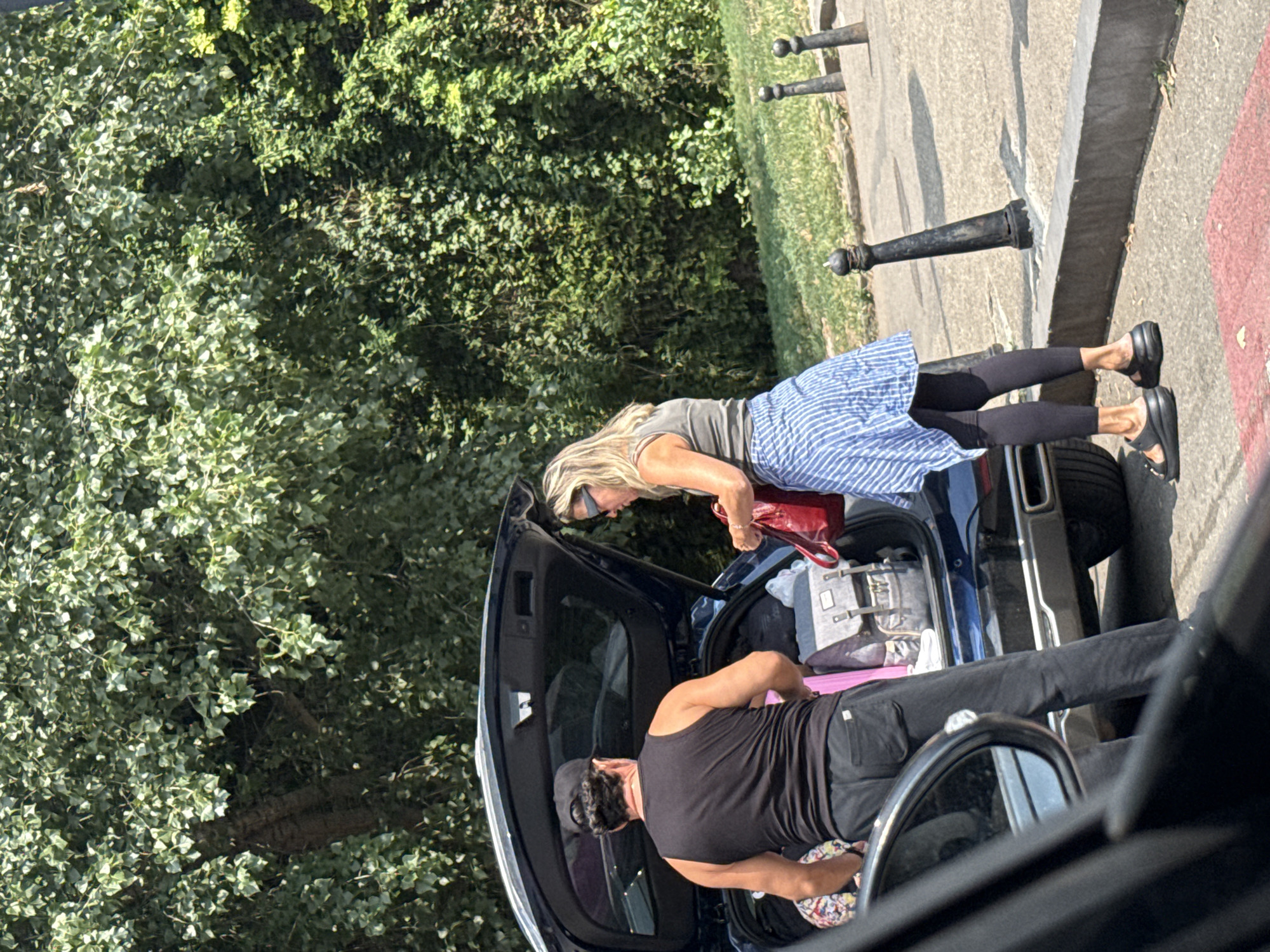 A man and a woman are standing by the open trunk of a dark blue SUV, packing it with luggage. The man, wearing a black tank top and black pants, is facing away from the camera. The woman, wearing a grey tank top, a blue and white striped skirt, and black leggings, is facing the trunk and holding a red handbag. The car is parked on a paved surface next to a sidewalk with decorative black bollards. Lush green trees and foliage form the background. The scene suggests preparation for travel or unloading after a trip.