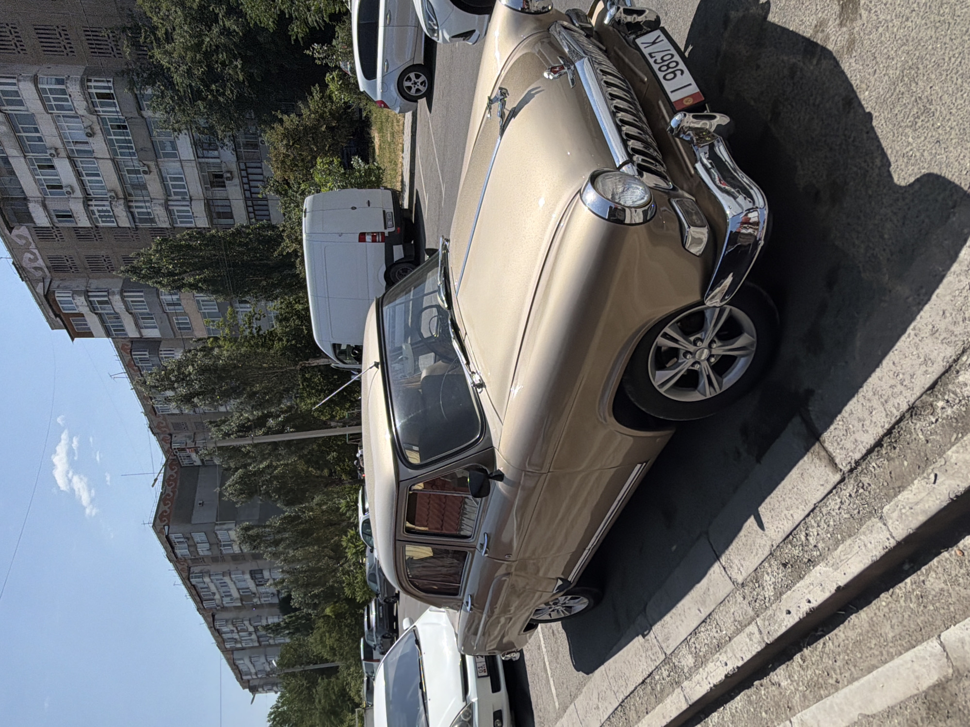 A vintage gold sedan is parked on a street in front of a multi-story apartment building. The car has chrome accents, a distinctive grille, and a hood ornament. A white van and other cars are visible in the background. The sky is blue with a few clouds. The scene appears to be a sunny day in a city.