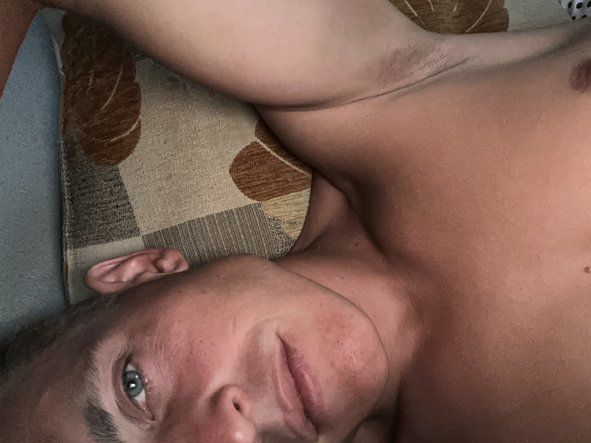 This is a close-up, overhead, indoor shot of a fair-skinned individual with short brown hair, lying down and looking towards the camera. The individual is shirtless, revealing their chest and armpit. They have blue eyes and are lying on a patterned surface that appears to be a bed or couch, with a blue and brown fabric. The lighting suggests it is daytime. There is no information in the image to indicate the location is Żary, Poland.