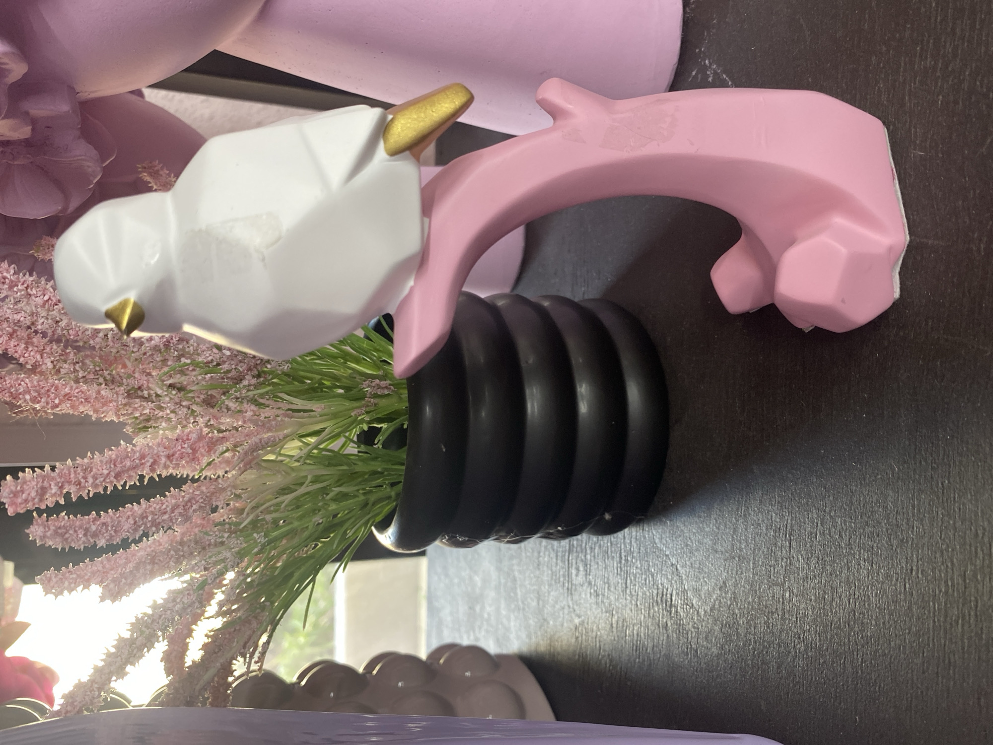 The image presents a close-up view of a decorative arrangement. A white, geometric bird figurine with a golden beak is positioned next to a pink, abstract sculpture resembling an arch or handle. Both elements are situated atop a black, ribbed vase filled with artificial flowers and greenery. The colors are muted, and the lighting appears to be soft and diffused. The location is not specified in the image. There is no indication this photo was taken in Yekaterinburg, Russia.
