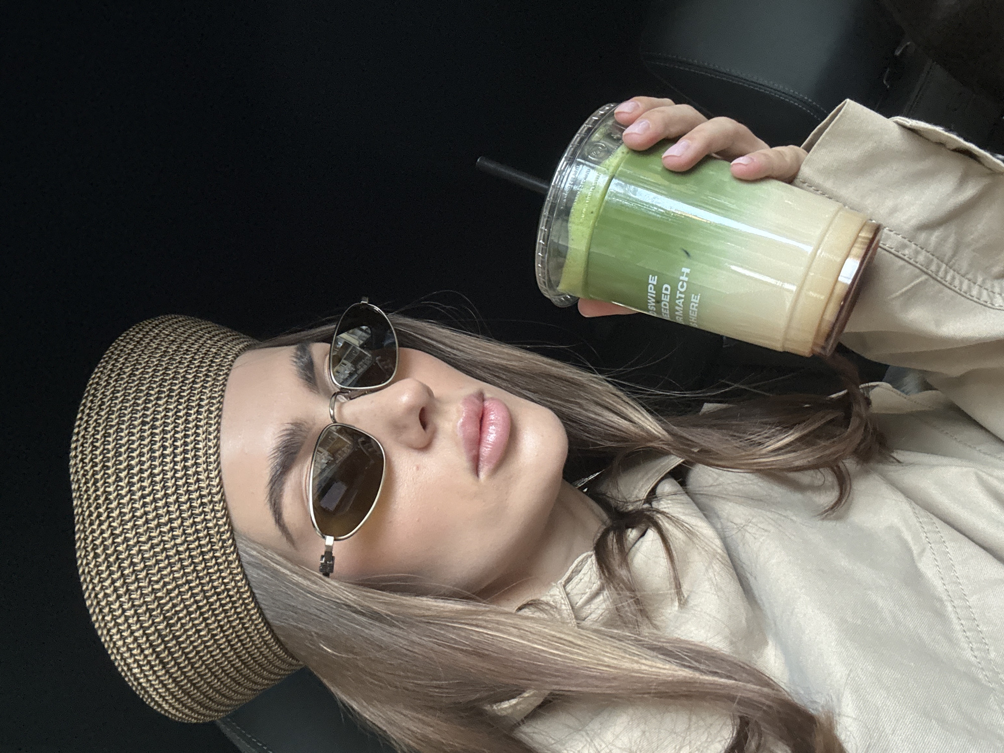 A woman poses indoors, holding a cup of matcha tea. She is wearing a tan beret and aviator sunglasses. She also wears a beige jacket. The woman is in a car, based on the black leather visible in the background. It is impossible to determine that the location is in the city of Warsaw, Poland based on the image provided.