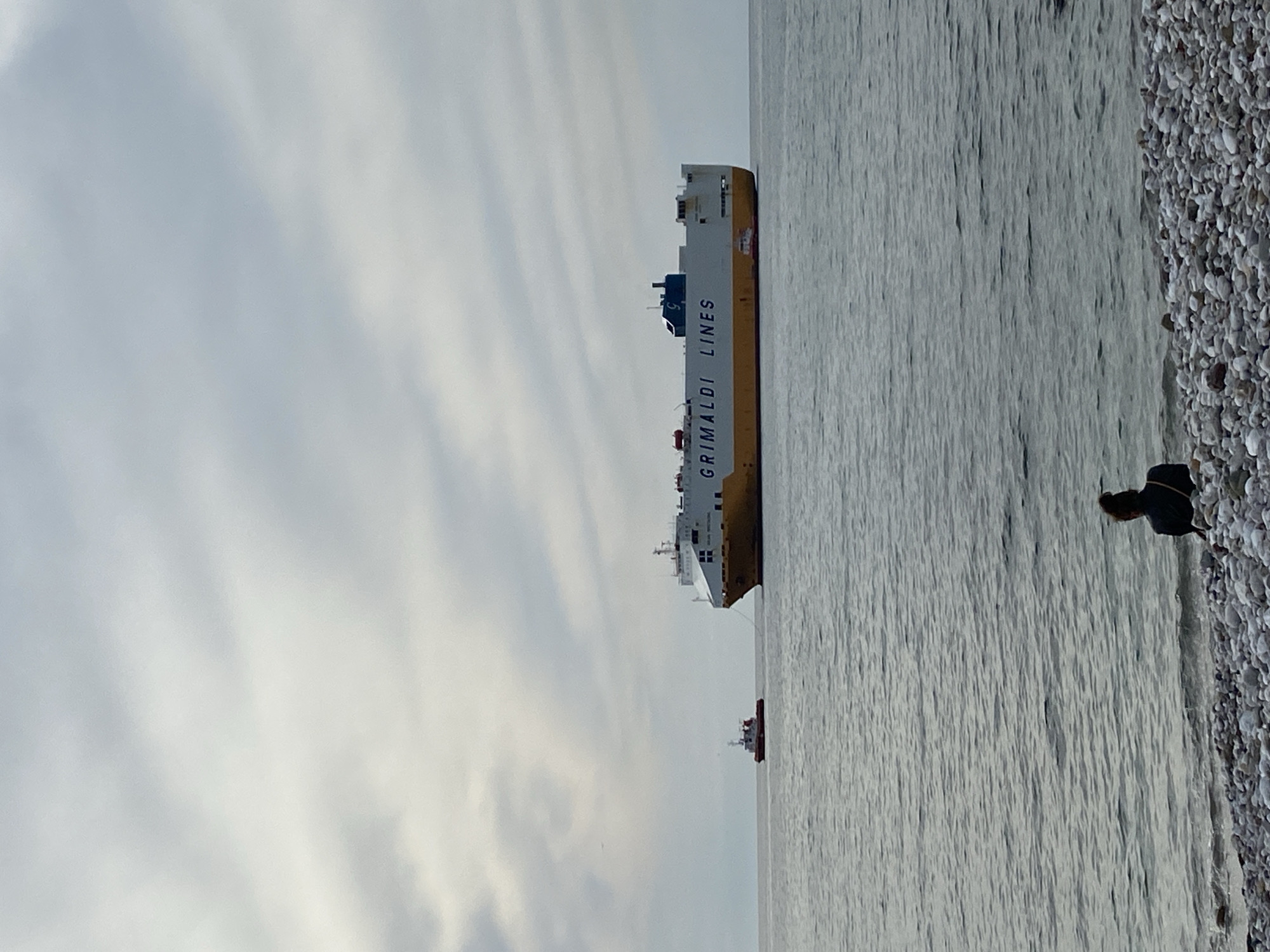 The image depicts a coastal scene in Bar, Montenegro. A large, yellow and white Grimaldi Lines cargo ship is positioned in the water, slightly off the shoreline. A smaller red and white vessel is also visible further away. The sky is overcast with a light-grey hue. Along the right edge of the image, a pebble beach slopes down towards the water. At the edge of the beach, a person is bending over, possibly examining something on the ground. The water appears calm with a slightly reflective surface.