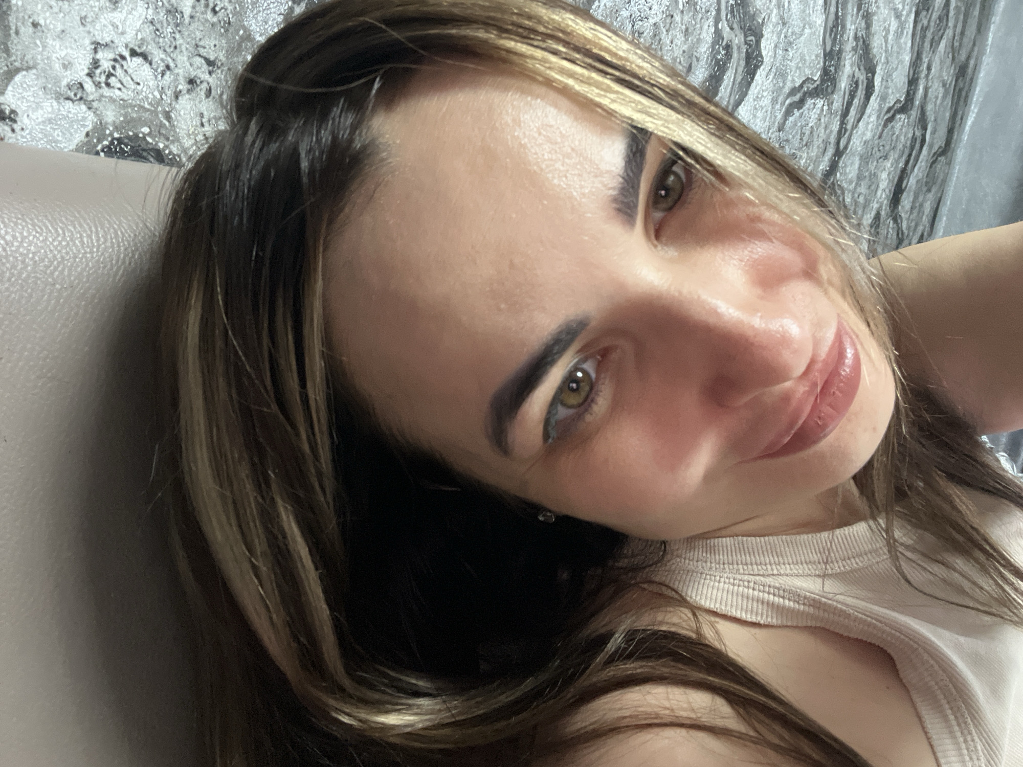 A woman is captured in a close-up, slightly tilted selfie, looking directly into the camera with a gentle, soft smile. Her face fills much of the frame, angled with her right side more prominent. She has captivating green or hazel eyes and well-defined dark eyebrows. Her lips are slightly parted, revealing a light pink, glossy finish. Her long hair, a blend of dark brown and prominent blonde or caramel highlights, frames her face and falls around her shoulders.
She is wearing a light-colored, possibly beige or off-white, ribbed top or tank top, which is visible at the bottom right of the image. The background elements suggest an indoor setting. To the left of her head, there is a smooth, light brown or beige upholstered surface, possibly part of a couch or headboard. Behind her and extending to the top right, a distinct textured wall or wallpaper with a silver and grey abstract, almost metallic-like pattern, is visible. The lighting appears to be indoors, and the woman's expression conveys a relaxed and amiable mood. There is no visible text in the image.
