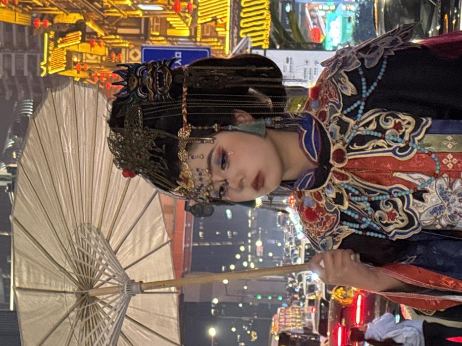 An individual with East Asian features is depicted from the chest up, positioned slightly angled to the left while looking forward. The setting is a bustling city street at night in Yuzhong District, China.
The person wears elaborate traditional-style attire. Their face features intricate makeup, including iridescent blue and orange eyeshadow, winged eyeliner, red lipstick, and a red teardrop design beneath the left eye. Their dark hair is styled into an ornate updo, adorned with numerous gold and green metallic embellishments, a prominent gold-colored forehead piece, and long dangling earrings with teal tassels. A dark, stylized mask-like ornament is affixed to the right side of the headpiece. The garment visible is a multi-layered traditional-style coat, predominantly black, with a richly embroidered red and blue collar and shoulders featuring intricate patterns and metallic thread accents. Strings of small, light blue beads are draped over the shoulders. The individual holds a light-colored, open parasol with a wooden handle in their right hand, extended upwards and to the left.
The background on the left shows heavy vehicular traffic, indicated by numerous red taillights and some white headlights, suggesting a road or bridge. Distant city buildings with illuminated windows are also visible. A partially obscured person in a light top and backpack is visible on the far left. The background on the right features a brightly illuminated building with traditional Chinese architectural elements and multiple red lanterns, characteristic of tourist or cultural areas. A blue sign with white Chinese characters, likely "入口" (rùkǒu, meaning "entrance"), is visible. The scene is illuminated by artificial streetlights, vehicle lights, and building lights.