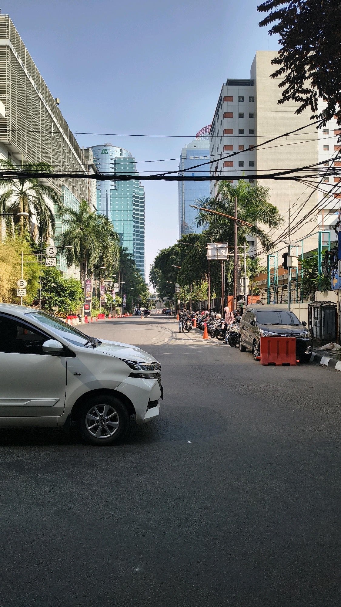 The image depicts a street scene in Kebayoran Baru, South Jakarta, Indonesia, on a clear day. Tall modern buildings, including a distinctive blue glass skyscraper, line the street. A white car is prominently featured in the foreground on the left, with its front-left wheel and bumper visible. On the right side of the road, a row of parked motorcycles is lined up along the sidewalk, some with riders present. A dark-colored SUV is parked behind the motorcycles, partially obscured by red traffic barriers. Palm trees and other foliage are interspersed between the buildings and along the street. Traffic signals and various signs are visible, indicating an urban environment. The road is paved, and there is a clear distinction between the road surface and the sidewalk. The sky is bright blue with no visible clouds.