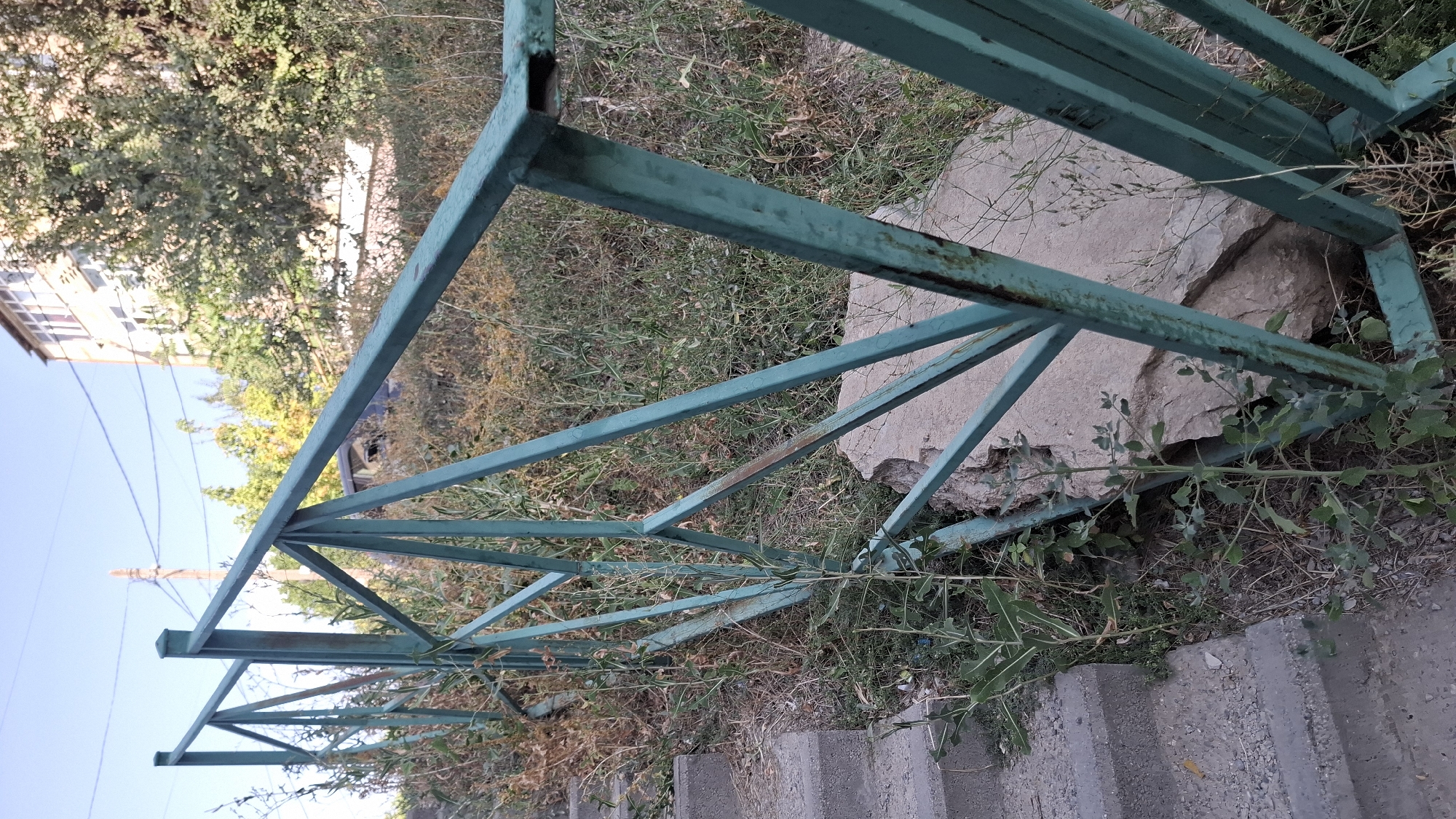An outdoor, slightly elevated, eye-level, medium shot captures a teal-colored metal fence with a geometric design. The fence runs diagonally from the lower left to the upper right corner of the frame. To the left of the fence are concrete stairs, some of which are visible. Beyond the fence, the ground is covered with dry grass and some green vegetation. In the background, a light brown building with white-trimmed windows is visible, partially obscured by trees with green and yellow foliage. Electrical wires and a utility pole are present in the sky. The scene is set during daylight hours, under a clear blue sky, with sunlight illuminating the environment. The overall impression is of an urban, possibly neglected, outdoor area.