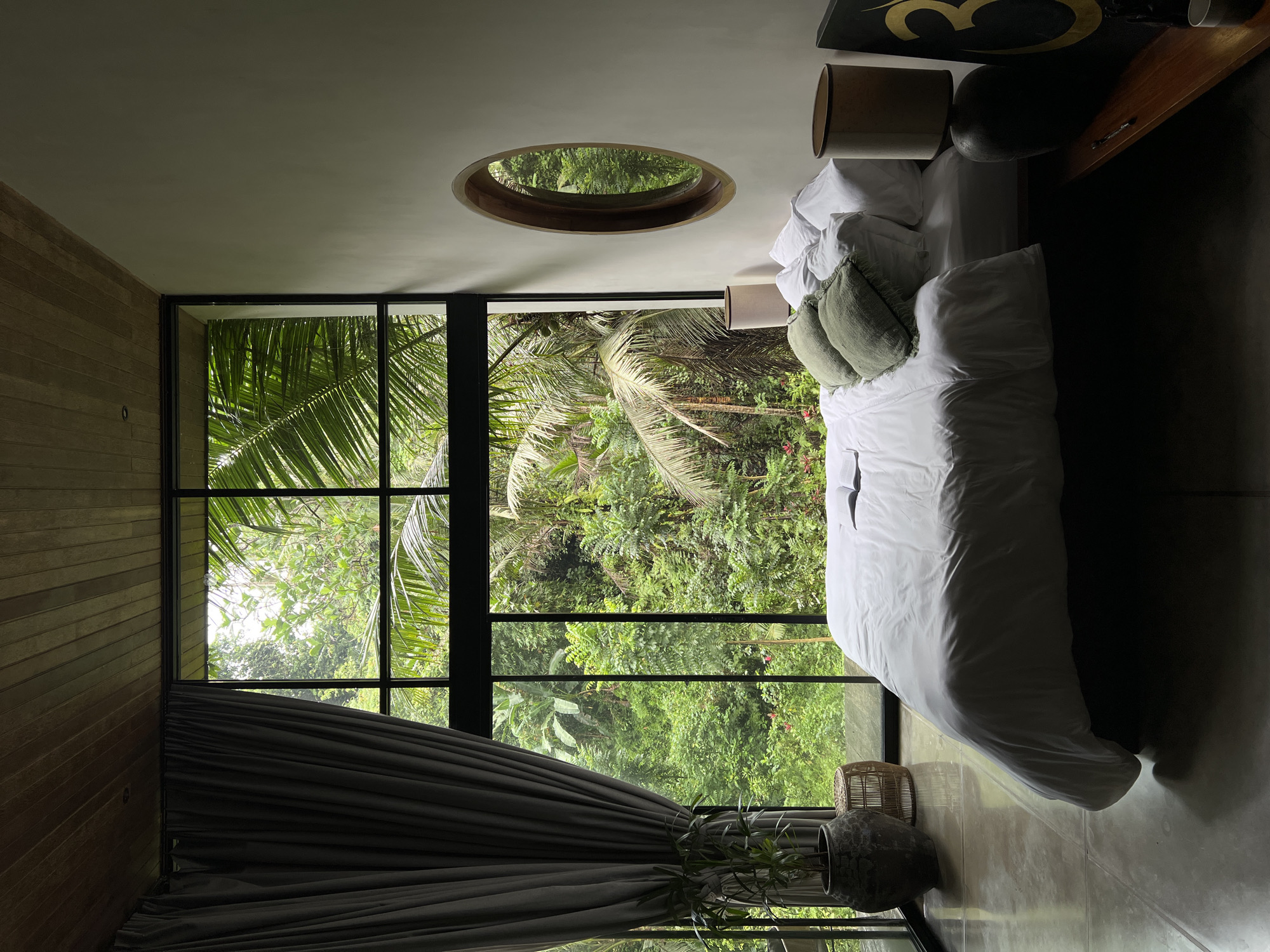 A bedroom in Ubud, Indonesia, features a large window overlooking a lush jungle. A white duvet covers a bed with several pillows, some of which are a muted green. A bedside table holds a lamp and a framed artwork. A round mirror hangs on the wall opposite the bed. A potted plant sits near the window, and a woven stool is placed beside it. The room has a concrete floor and a wooden ceiling.