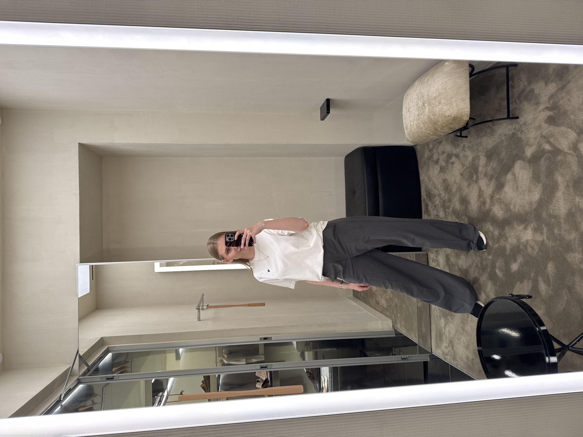The image displays a woman taking a mirror selfie within a well-lit, neutral-toned interior, likely a changing room or boutique in a retail establishment. The specific city of Perm, Russia, cannot be discerned from the visual information alone.
The woman, with blonde hair, is positioned centrally, holding a dark-colored smartphone in her right hand to capture her reflection. She wears a white short-sleeved t-shirt with a small dark emblem on the left chest, dark gray wide-leg trousers, and black and white athletic shoes. A metallic bracelet adorns her left wrist, and a thin silver necklace with a small pendant is visible.
The full-length mirror, flanked by vertical LED light strips, reflects the room. To the left of the woman's reflection is a glass-fronted wardrobe or display cabinet containing shoes (brown loafers, beige boots) and folded clothing on shelves. Above the wardrobe, a small section of a door or panel is slightly ajar. A silver metal rail with two wooden hangers extends from the wall near the wardrobe.
To the right of the woman's reflection, a small black floating shelf is mounted on the wall. A rectangular black upholstered bench with a buttoned top is situated against the wall to the woman's left. Further to the right in the foreground, a smaller rectangular stool with a light beige, textured cushion and a black metal frame is present. In the left foreground, a round, glossy black table with a tripod base and a ring handle sits on the floor.
The walls are covered in a light, vertically ribbed textured wallpaper or paneling. The floor is covered with a muted gray or brown textured carpet. Recessed lighting is visible in the ceiling reflected in the mirror. The overall environment suggests a modern, organized commercial space.