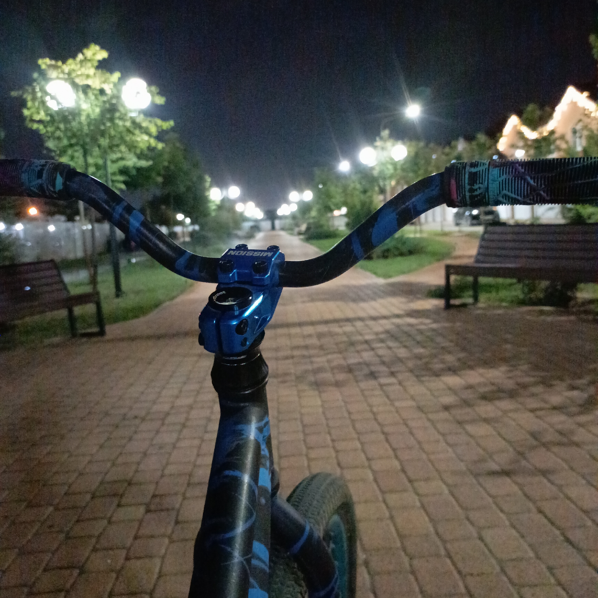 The image is a first-person perspective shot from a bicycle at night. The bicycle's handlebars and stem are prominently in the foreground, with a blue stem bearing the "MISSION" logo. The handlebars have black grips with blue patterns. The bicycle is on a paved walkway made of interlocking rectangular stones. In the background, trees with illuminated streetlights are visible, creating bokeh effects. There are benches along the walkway, and a building with decorative lights can be seen in the distance. The scene depicts a park or public area in the city of Krasnodar, Russia, likely in the evening or at night, given the artificial lighting and dark sky. No people are visible in the image.