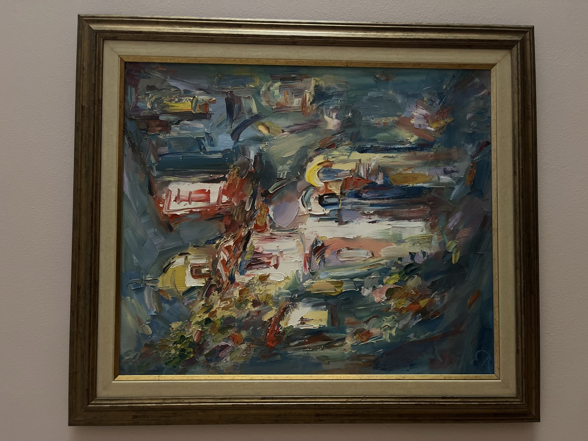 An abstract or semi-abstract oil painting, characterized by thick impasto and bold, visible brushstrokes, is displayed in a wooden frame against a light wall. The painting's subject appears to be an urban or village landscape, possibly depicting a jumbled arrangement of buildings, roofs, and streets, rendered with dynamic forms and vibrant colors.
The palette is dominated by cool blues, grays, and deep greens, which form the background and give a sense of depth or sky/water elements. These are contrasted sharply with warmer tones of white, cream, vibrant yellows, reds, and oranges, which define what seem to be architectural structures or prominent ground features. The heavy application of paint creates significant texture across the canvas, making individual brushstrokes and layers clearly visible. There are no discernible human figures or explicit activities taking place, as the focus is on the fragmented landscape.
Notable details include the painting's frame, which is a two-part design: an inner, lighter-toned (possibly cream or light wood) liner and a more ornate outer frame with a gilded or dark bronze finish and a subtly textured or patterned surface. The overall impression is one of movement and energy conveyed through the artist's expressive style. In the bottom right corner of the painting, there is a small, stylized signature or monogram, which is difficult to decipher but appears to be a combination of letters, possibly an "R" or "B" intertwined with another character. The ambient lighting suggests an indoor setting, with the painting illuminated to showcase its textures and colors.