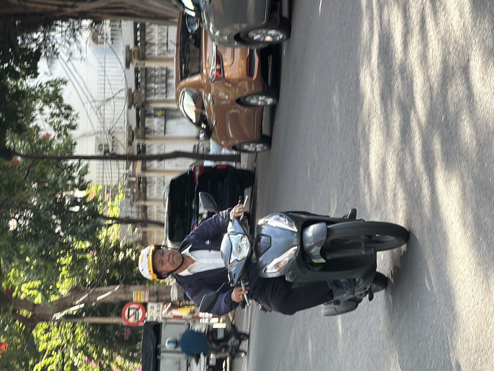 A middle-aged man wearing a white collared shirt, a dark blue jacket, and a yellow and white helmet rides a dark gray scooter down a paved city street. He is wearing dark pants and dark shoes, and has rings on two fingers of his right hand. The scooter has a dark gray body with silver accents and two mirrors. In the background, a brown sedan is parked behind a dark SUV, and a lighter gray car is parked to the right of the brown sedan. A white building with ornate fencing is visible behind the cars, with a tree on the right side of the image. A traffic sign with a red circle and a diagonal line through a motorcycle symbol is visible on the left side of the image, along with a white truck and another scooter. The street is lined with trees, casting shadows on the asphalt. The scene appears to be in Nha Trang, Vietnam, given the presence of scooters and the style of the buildings and traffic signs.