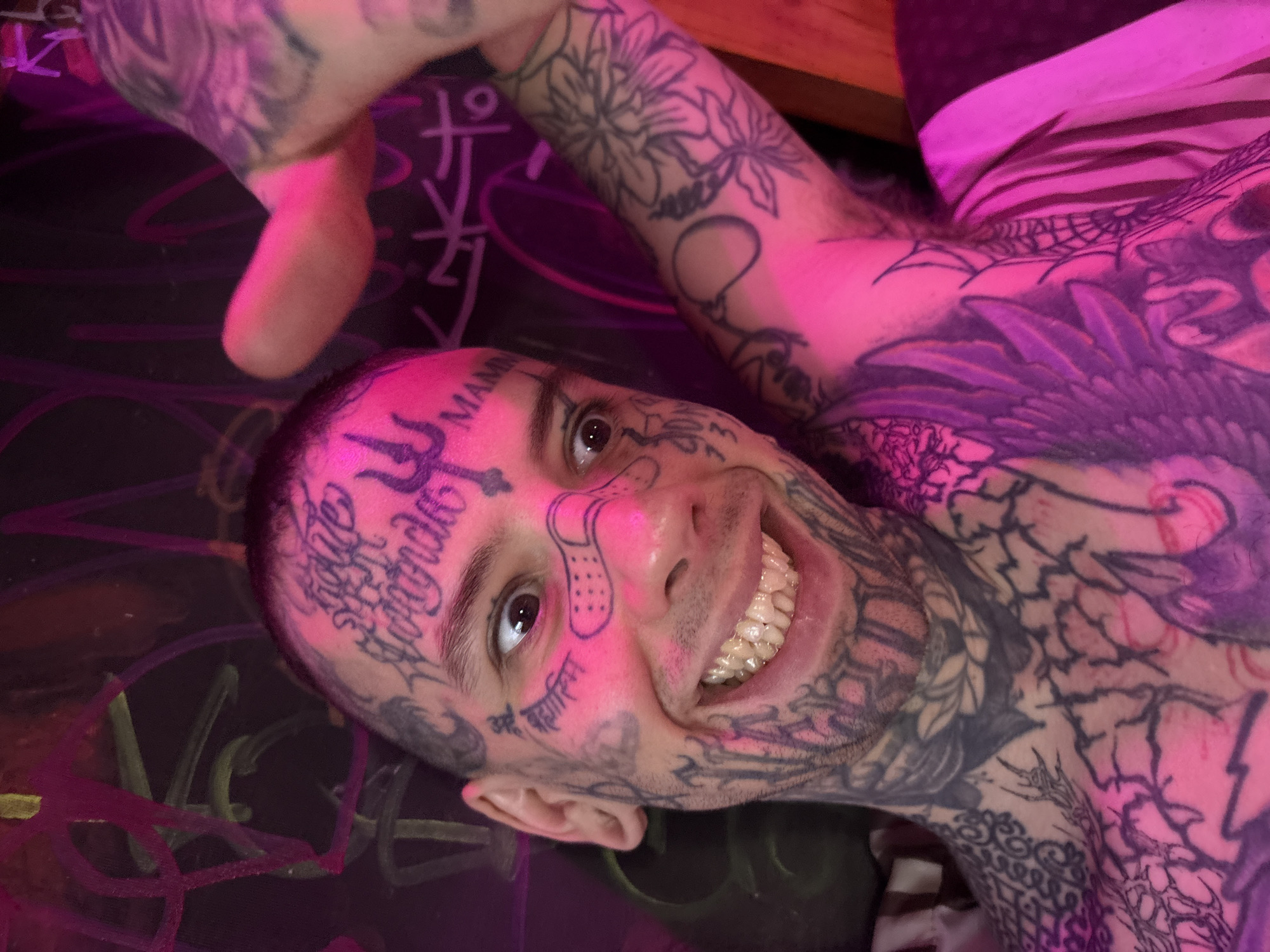 A man with extensive face and body tattoos is shown close up. The tattoos include various designs and lettering in different scripts. He is smiling, revealing his teeth. A bandage tattoo is on his face. He is lying on a surface with dark colors and graffiti-style drawings. The lighting is primarily pink. The location in Buenos Aires, Argentina, cannot be determined from the image.