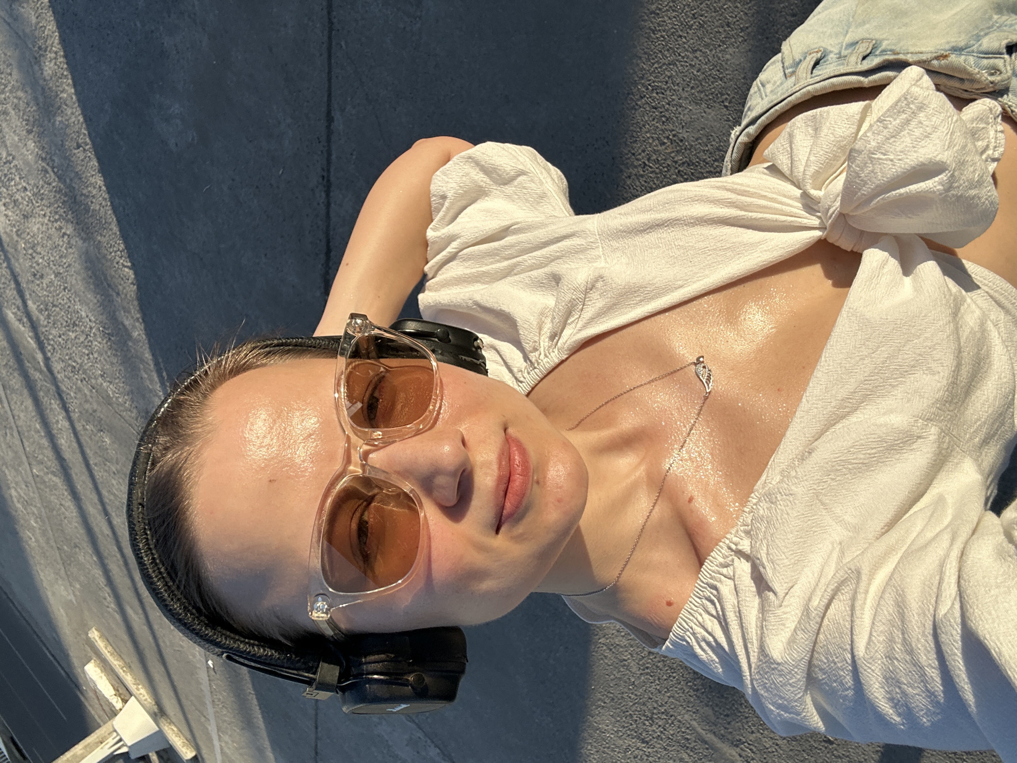 In Baan Hua Thanon, Thailand, a woman is captured in a selfie during golden hour. She wears black headphones, stylish clear-rimmed sunglasses with amber lenses, and a white tie-front blouse. A delicate silver necklace with a wing pendant rests on her chest, which glistens with sweat. The background is a rough, gray paved surface, partially shaded by a long, diagonal shadow. The overall lighting suggests an outdoor setting with warm sunlight.