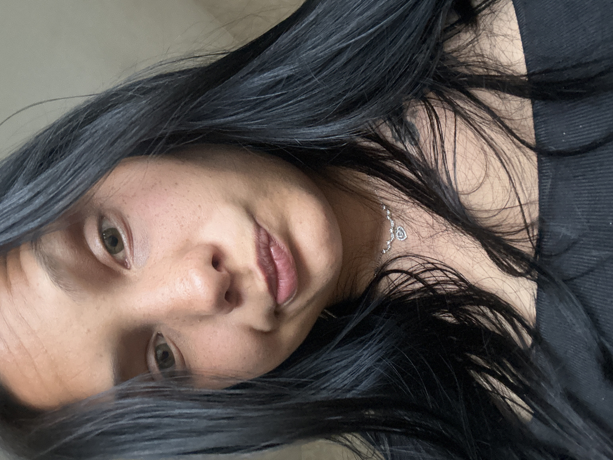 A close-up, tilted shot shows a woman with dark hair, light skin, and green eyes. She is wearing a necklace with a heart pendant and a black top. The background is indistinct, but appears to be a room with light-colored walls. There is no indication of Sofia, Bulgaria, or any other specific location.