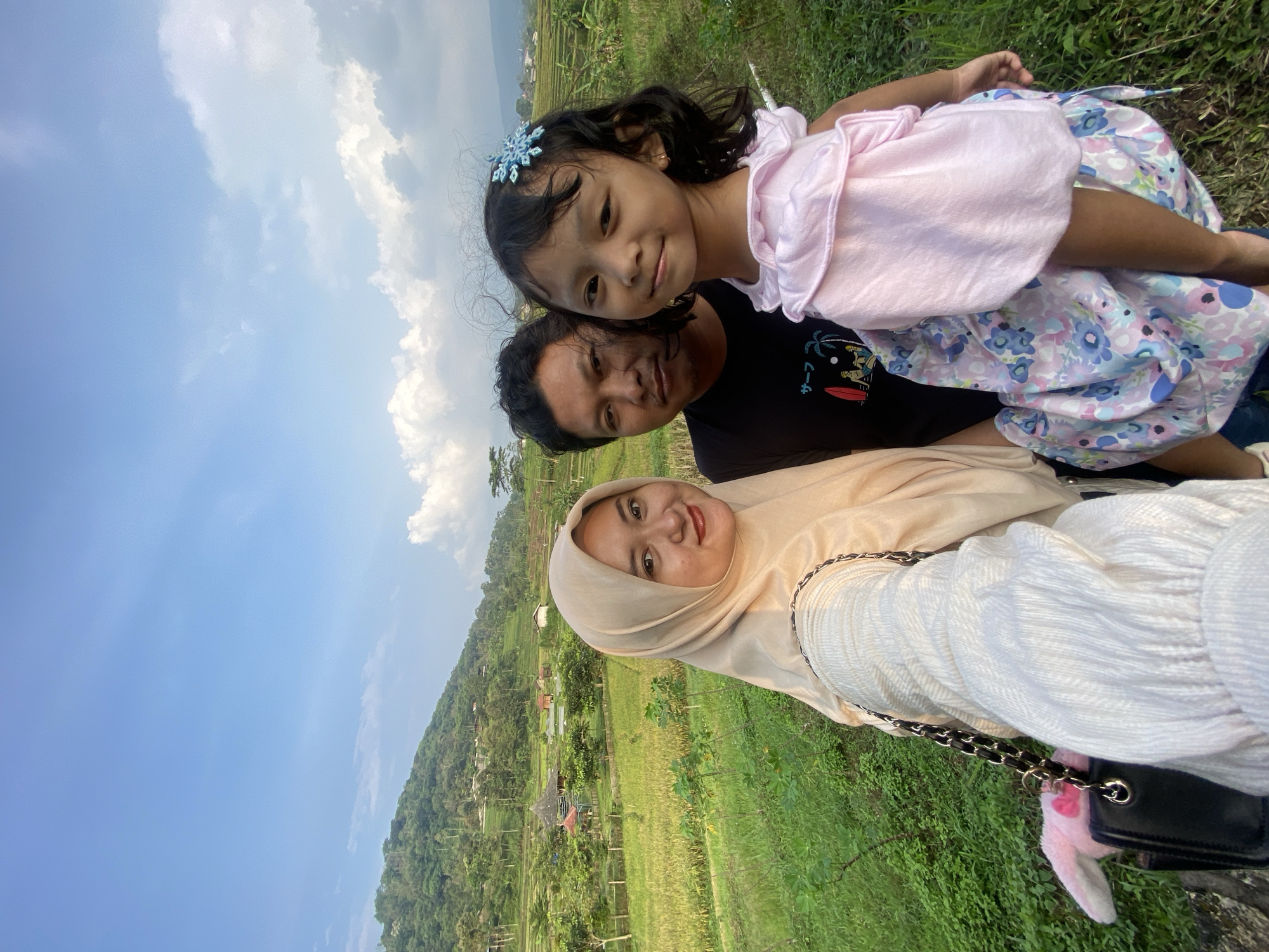 A family portrait of three people is taken outdoors. A woman wearing a hijab stands in the foreground. Behind her is a man, and a young girl is in the back. The little girl is wearing a light pink dress with a blue flower on her hair. They are on a grassy terrain that appears to be near farmland. The background features a landscape with trees, rice fields, and houses. The sky is blue with some clouds. The location appears to be Penanggungan, Indonesia.