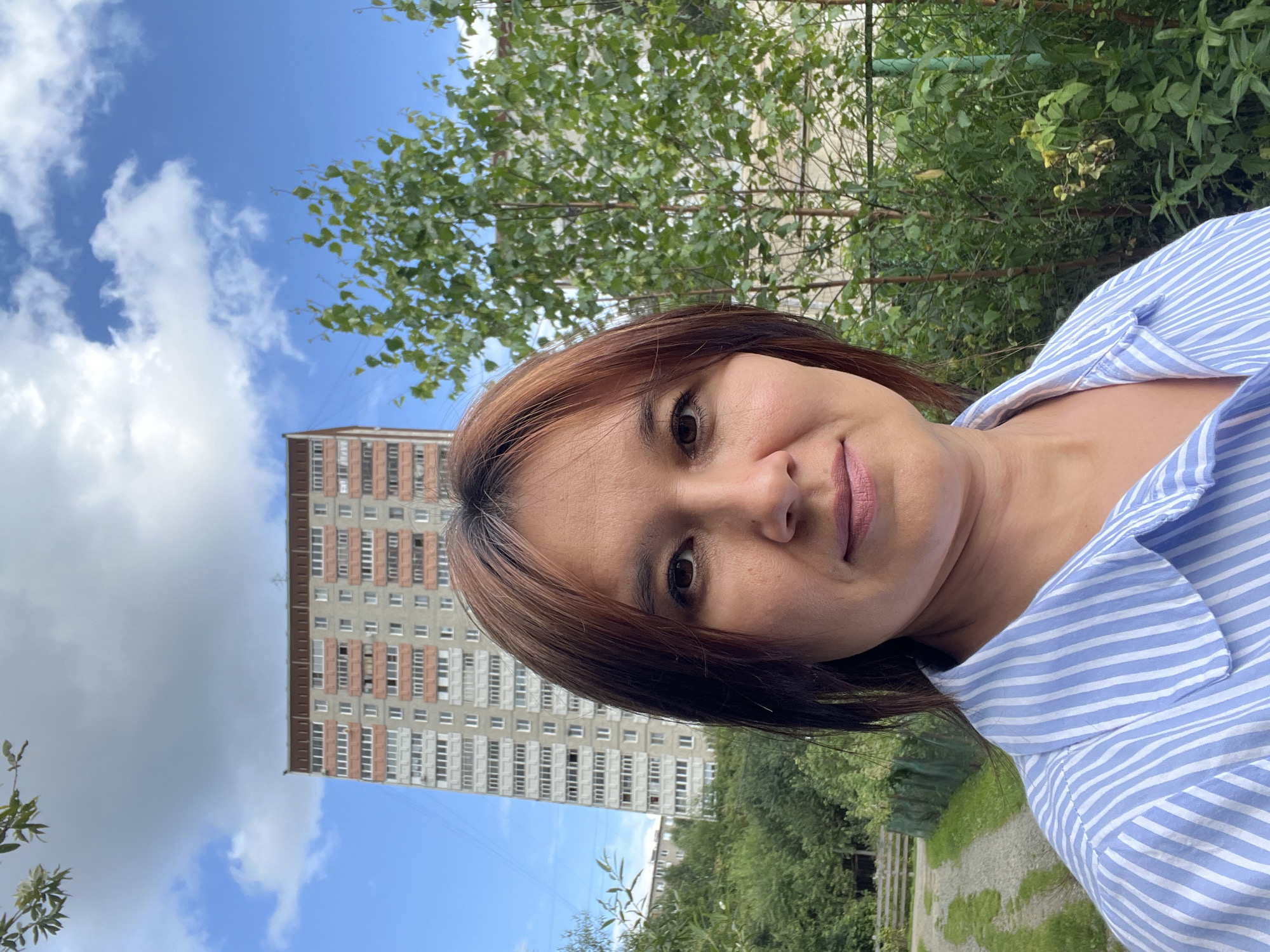 A selfie shows a woman with medium-length brown hair, parted in the middle, with reddish undertones. She has brown eyes and is wearing a blue and white striped button-down shirt. She is standing outside with a tall, multi-story apartment building in the background, characterized by its pink and grey facade and numerous windows. To the right of the building, a leafy green tree is visible, and further to the right, a green fence can be seen. The sky above is blue with scattered white clouds. The ground in front of the woman appears to be a gravel path leading past a green fence and more foliage, suggesting an urban or suburban setting. The presence of the apartment building and the style of architecture are consistent with residential areas in Pervouralsk, Russia.