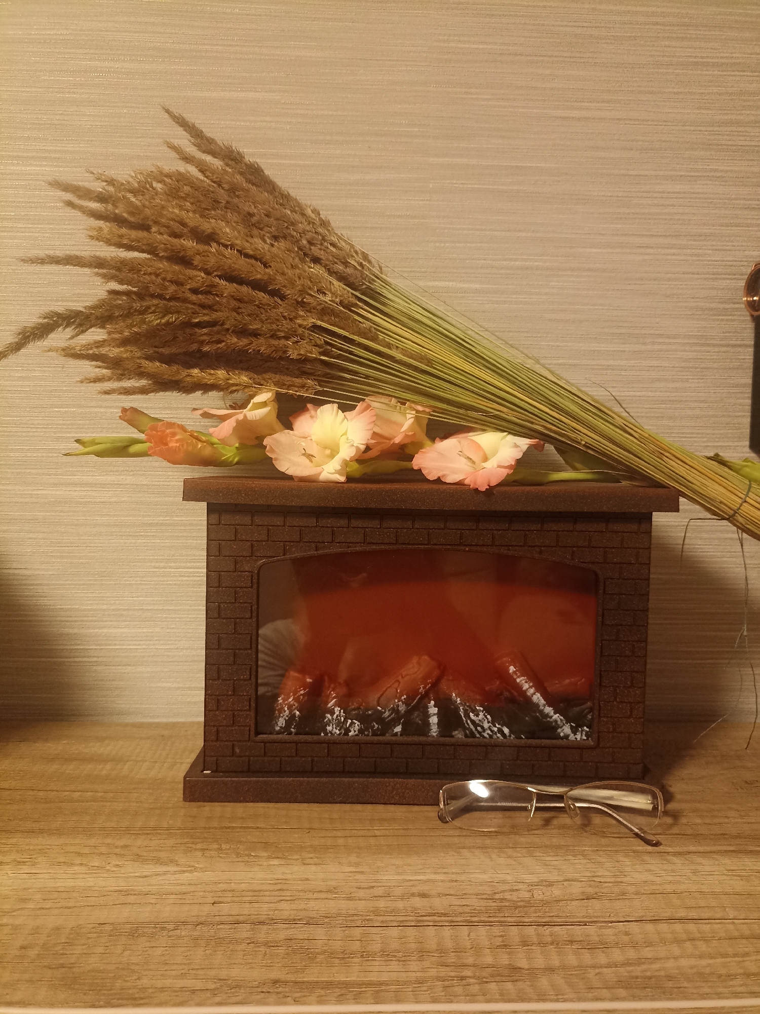 A bouquet of dried grass and light pink gladiolus flowers rests across the top of a miniature brown brick fireplace. The fireplace has a faux-flame insert glowing orange and red. In front of the fireplace, on a wooden surface, sits a pair of thin-rimmed eyeglasses. The background is a textured, light beige wall. The scene is likely indoors, on a shelf or table, with no discernible city or geographical features of Homyel, Belarus.
