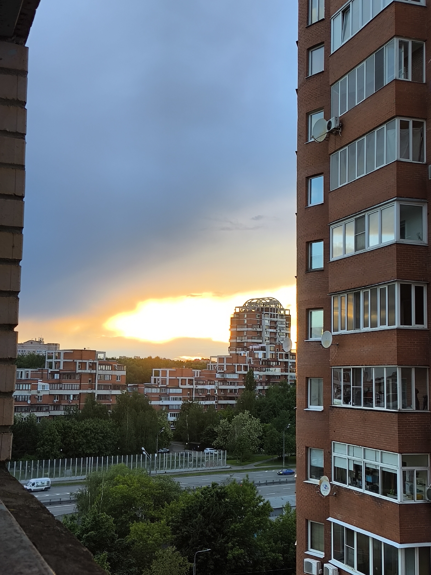 View of residential buildings from a window, with a sunset sky in the background. The buildings are made of red brick. A highway is visible in the middle ground. The sky shows shades of blue, gray, and orange from the sunset.