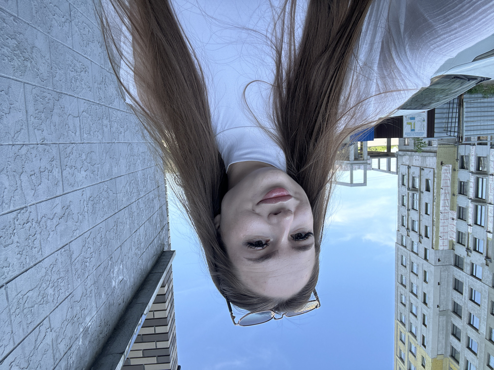 A young woman with long, brown hair and sunglasses perched on her head is taking a selfie outdoors. She is wearing a white turtleneck shirt. She is standing in front of a textured gray wall. To her left, there is a tall, unfinished apartment building and a white car parked. The sky is blue with a few clouds. The scene appears to be in a construction or urban area.