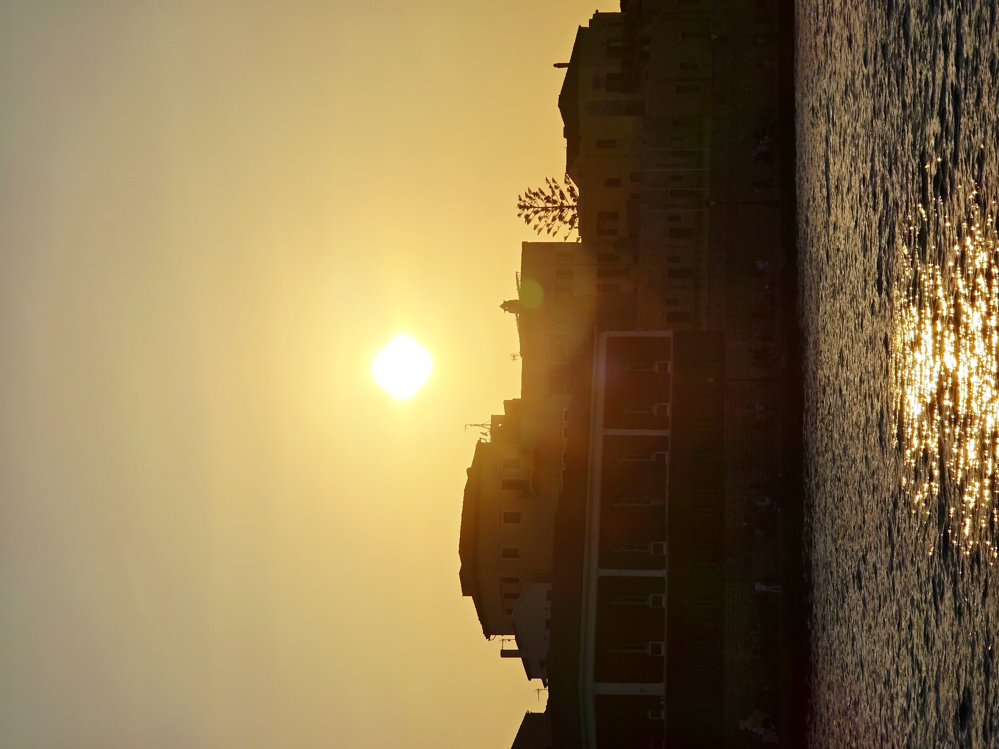 This image depicts a sunset over the city of Chania, Greece, specifically showcasing the Venetian Harbor. The sun, a bright, luminous orb, is positioned in the upper center of the frame, casting a warm, golden glow across the sky and the water. Silhouetted against this vibrant sky are the distinct architectural features of buildings lining the harbor. These structures exhibit a variety of rooflines and window placements, with some featuring colonial-style columns in the foreground. The water in the foreground is textured with ripples, reflecting the sunlight in a glittering pattern, suggesting a calm sea. A scattering of people, appearing as small, dark figures, are visible along the waterfront, some appearing to be walking or standing, while others are seated. A distinctive, slender tree with a feathery canopy stands out against the skyline, adding a unique natural element to the urban landscape. The overall impression is one of a serene evening scene in a historic coastal city.