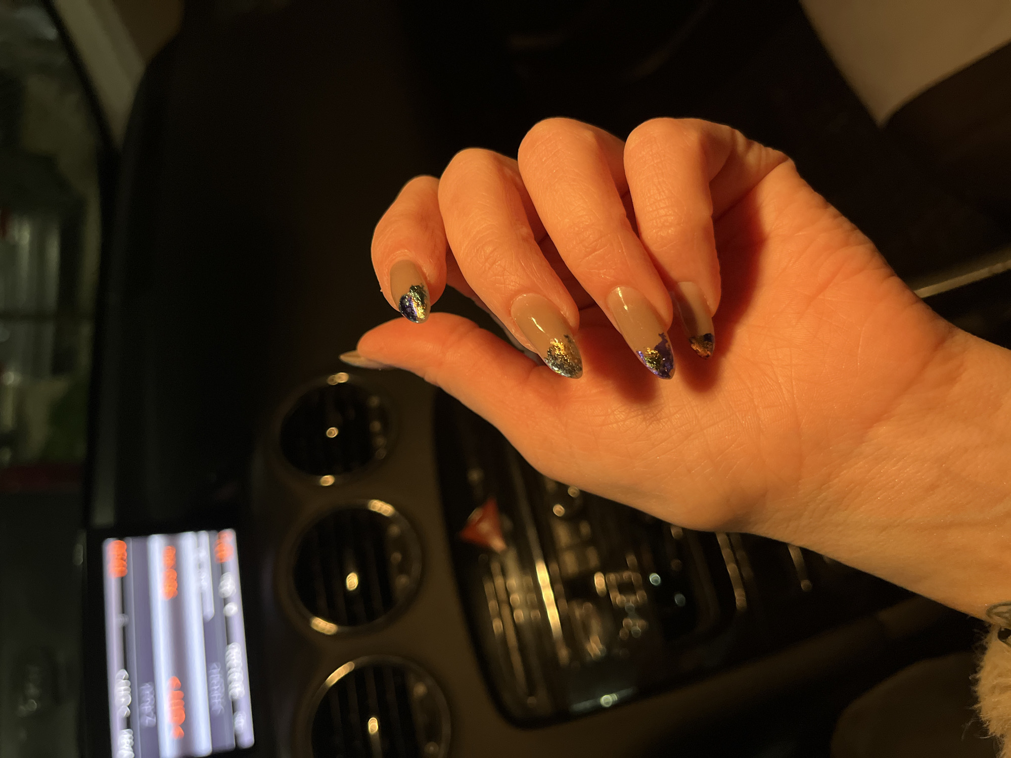 A human hand with painted nails is positioned in front of a car dashboard. The nails are painted with a beige color and have a decorative accent at the tips consisting of iridescent glitter. The hand is slightly curved, with the fingers bent. The car dashboard is dark, featuring circular air vents and a digital display screen that shows vertical lines of white and orange. The image appears to be taken in a dimly lit interior, possibly inside a car. There is no indication of the location being Varna, Bulgaria.