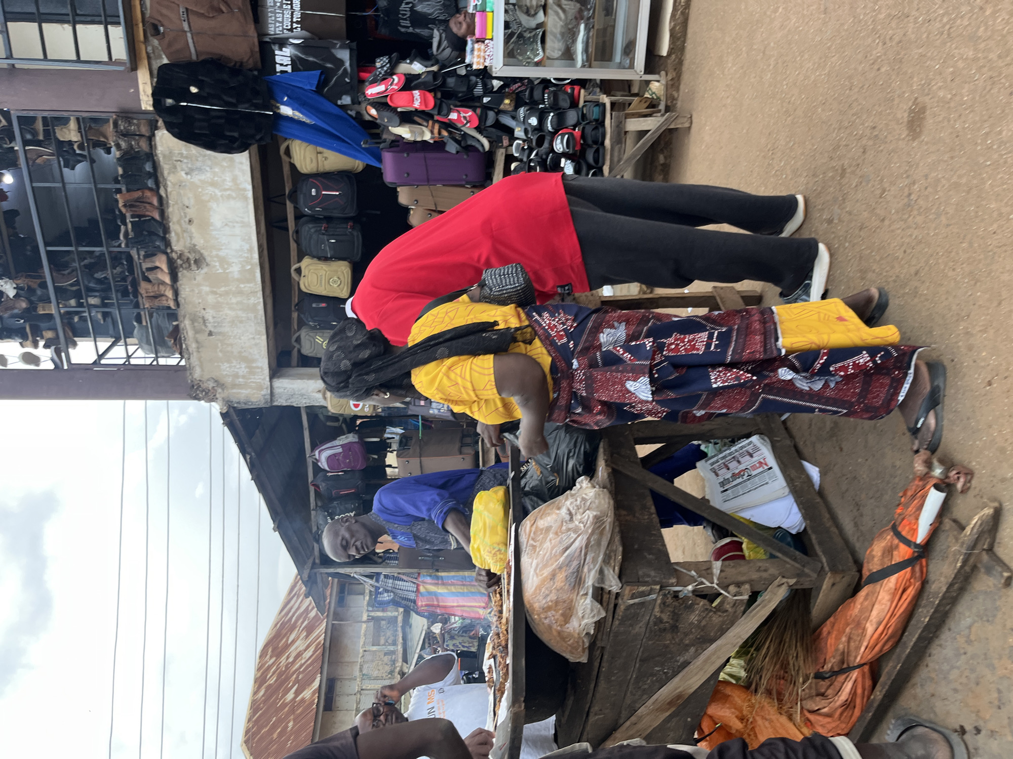 The image depicts an outdoor market scene in Bukuru, Nigeria, likely a food stall area. In the foreground, a man wearing a blue shirt and apron stands behind a wooden stall laden with food, including skewers of grilled meat. A woman in a vibrant yellow and patterned skirt stands to his right, facing away from the camera. To the left, a person in a white t-shirt, presumably speaking on a phone, is also near the stall. Behind the stall, various items are displayed, including bags and what appear to be shoes or boots on shelves in the background buildings. The environment suggests a bustling commercial area with a variety of goods for sale.