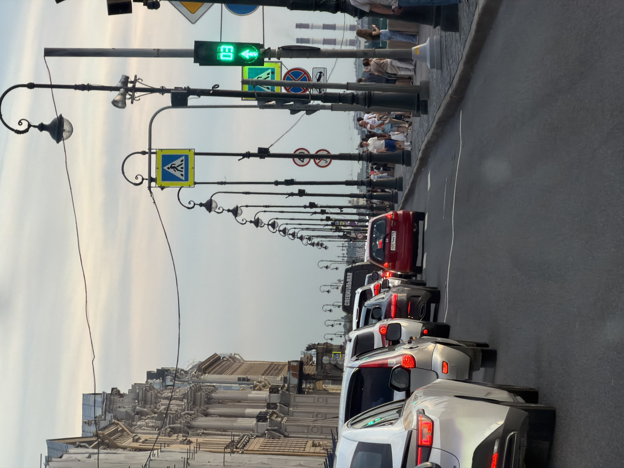 The image depicts a traffic jam on a bridge in Saint Petersburg, Russia, with a line of cars on the road. On the left, ornate buildings with classical architecture line the street. The sky is overcast with soft clouds. Several streetlights with decorative designs illuminate the scene. Pedestrian crossing signs and traffic signals are visible, with one traffic light displaying a green arrow and the number "03". A group of people are walking on the sidewalk. In the distance, industrial chimneys can be seen. The overall atmosphere suggests a typical urban setting during the daytime.