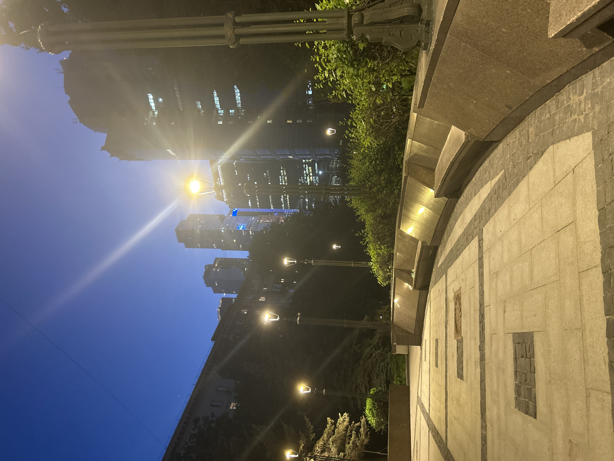 The image depicts an urban scene at night in an unknown city. In the background are several high-rise buildings with numerous illuminated windows, suggesting commercial or residential occupancy. A row of streetlights with glowing lamps lines the path towards the buildings. These lights cast a warm glow, contrasting with the cool blue twilight sky. The foreground features a stone structure with a curved edge, suggesting a wall or a section of a bridge, adorned with intricate designs and patterns. The overall atmosphere is calm, with the architectural details enhanced by the lighting, creating a sense of depth and urban sophistication. The location appears to be a bridge or elevated pathway providing a view of the city skyline.