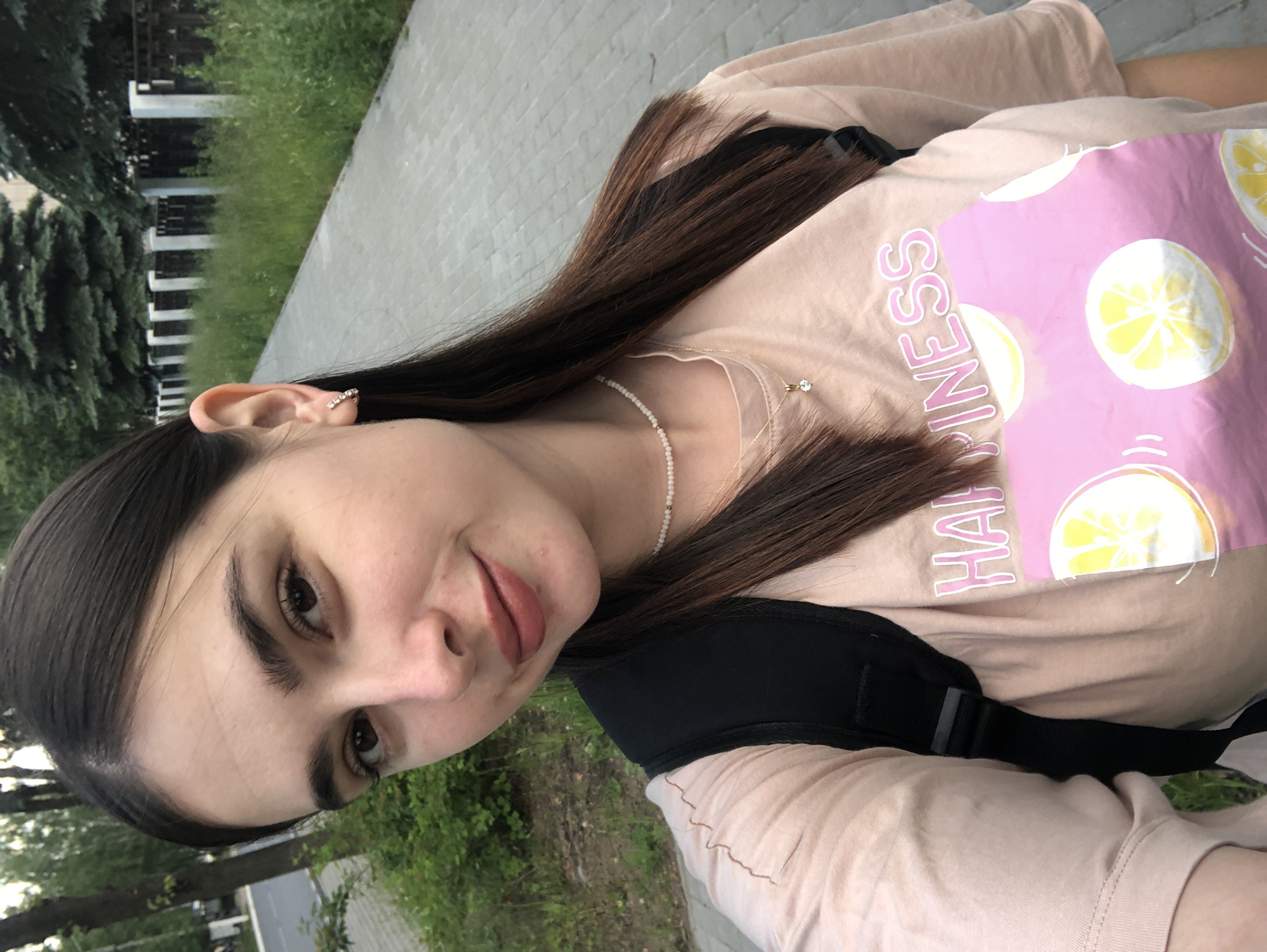 A young woman with dark brown hair, brown eyes, and fair skin is taking a selfie outdoors. She is wearing a light pink t-shirt with a graphic design of lemons and the word "HAPPINESS" printed on it. She also has on a short, beaded white necklace and a delicate gold pendant. A black backpack is visible over her shoulders. The background shows a paved walkway, green grass, trees, and a wooden fence with white pillars. The lighting suggests it is daytime. The scene is consistent with a park or a residential street in Chaikovsky, Russia.