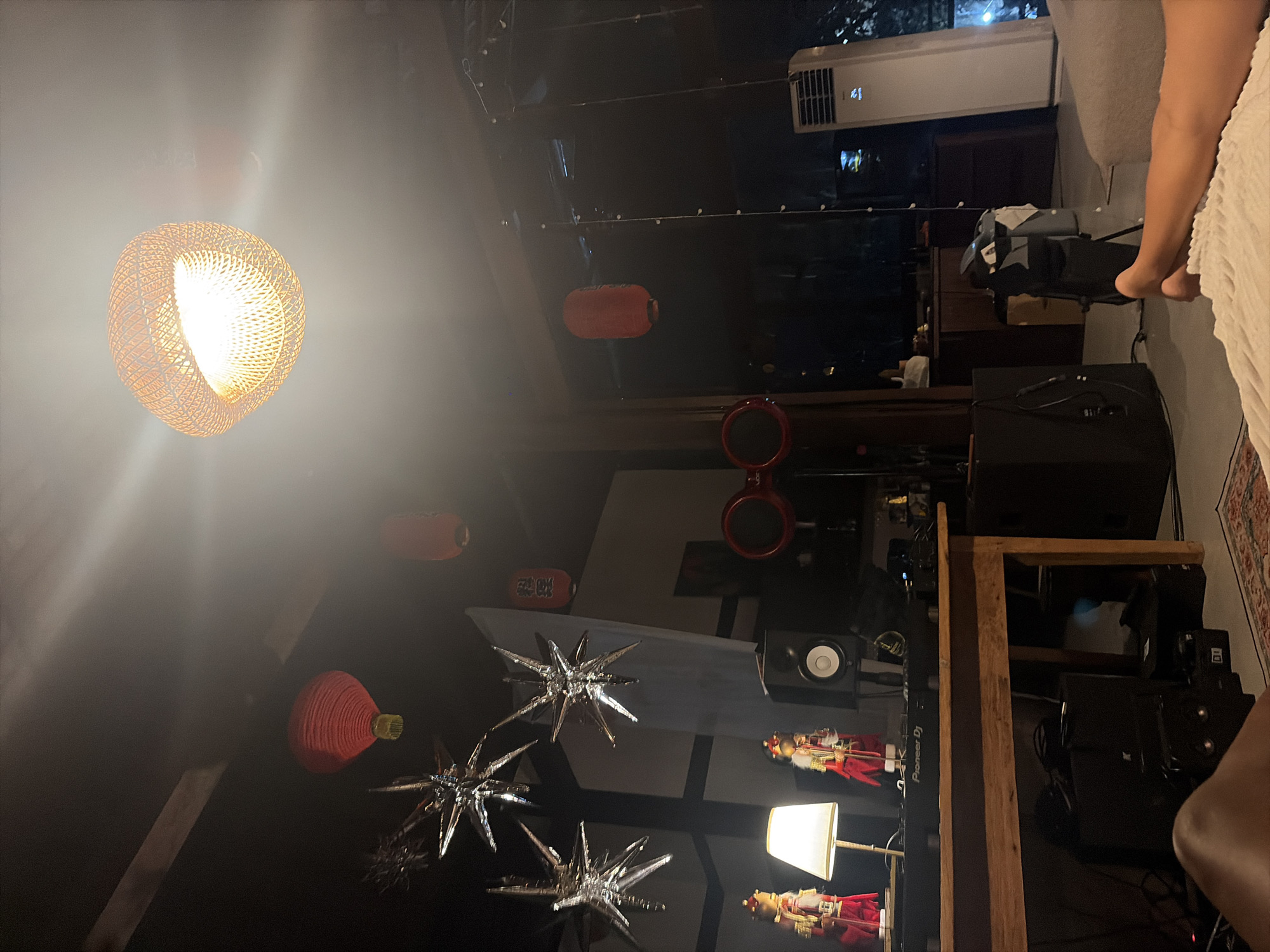 The image depicts an indoor scene, possibly a living room or entertainment space. In the foreground, a person's legs are visible, resting on a textured surface, suggesting they are lying down. The room is dimly lit, with several red lanterns hanging from the ceiling, adding a touch of Asian decor. A prominent woven lampshade hangs from the ceiling, emitting a warm glow.
In the mid-ground, a wooden table is set up with DJ equipment, including a "Pioneer DJ" controller. Two decorative figures, dressed in traditional attire, stand on the table. A white lamp illuminates the figures. Behind the DJ setup, a black speaker is positioned.
Further back, there are more speakers, including a large black one on the floor. A red, circular object, possibly headphones or a decorative piece, is mounted on a stand. Through a large window or glass partition, a dark exterior is visible, hinting at a nighttime setting. An air conditioning unit stands tall on the right side of the room. The overall ambiance suggests a relaxed, personal space with a focus on music and decor. There is no information in the image to suggest the location is in Kediri, Indonesia.