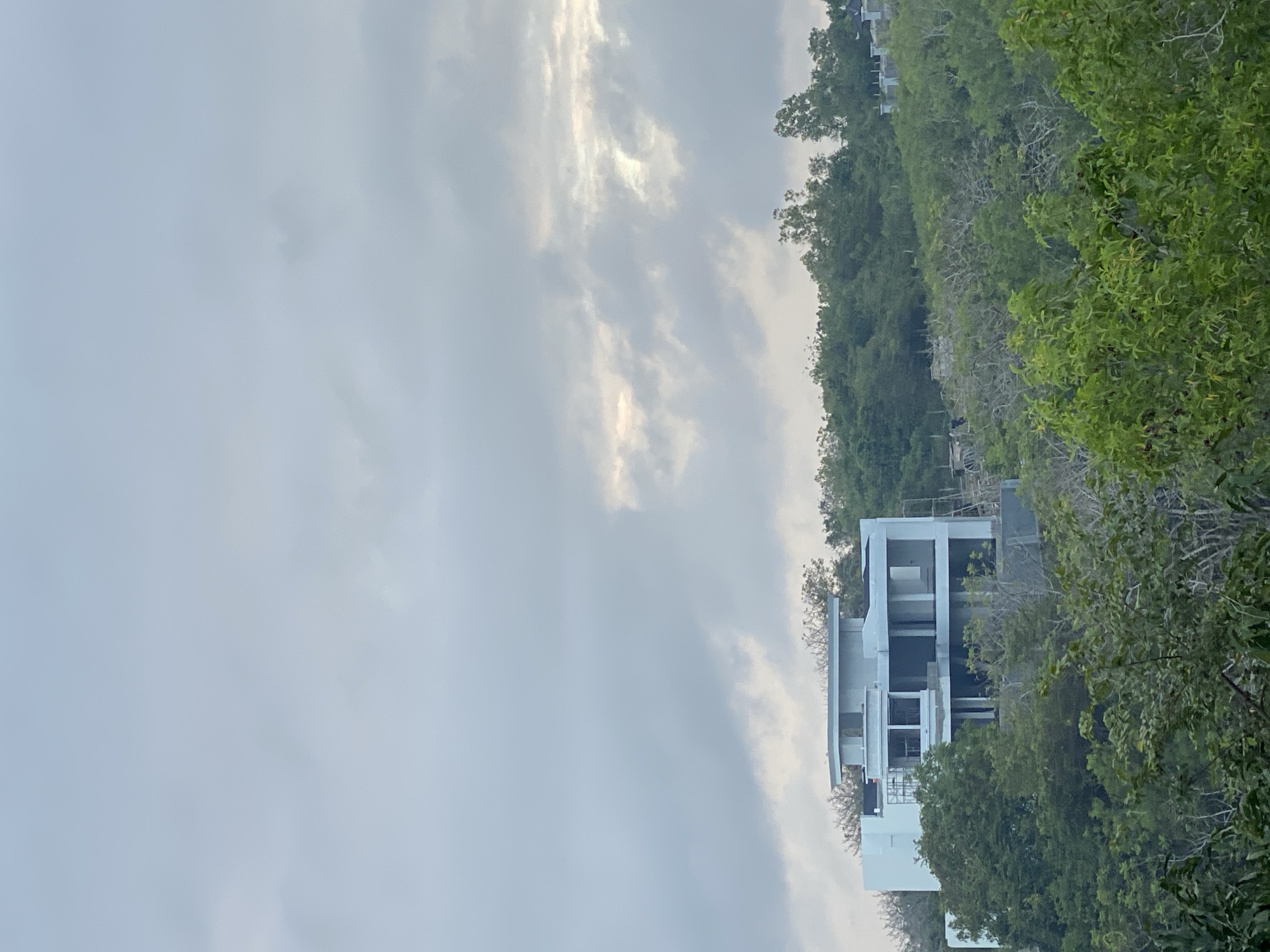 An outdoor, medium shot captures a modern, multi-story white house under construction, nestled amidst lush green trees on a hillside. The sky above is overcast with clouds, with a sliver of sunlight peeking through the diffused grey. The house features a geometric design with multiple balconies and open spaces, revealing the structural framework. In the foreground, dense foliage obscures parts of the lower sections of the hill and house. The overall impression is one of a developing residential area in a tropical or subtropical environment, likely Kuta Selatan, Indonesia, given the vegetation and architectural style. There are no people or discernible actions within the frame.