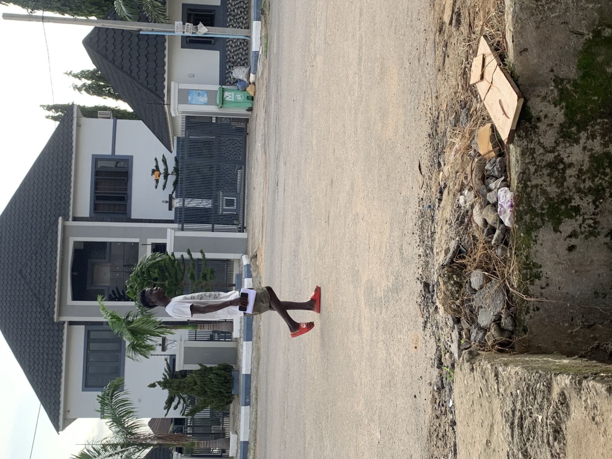 A young man in a white t-shirt and green shorts walks across a paved street in Abuja, Nigeria, holding a booklet. He is wearing red sandals. In the background, a modern, multi-story house with a dark roof and gray exterior is visible, surrounded by a fence and several trees, including palm trees. A green garbage bin is positioned near the entrance to the property. The street in the foreground is worn and has a drainage ditch filled with debris, including rocks, dried grass, and a piece of wood. The sky is overcast.