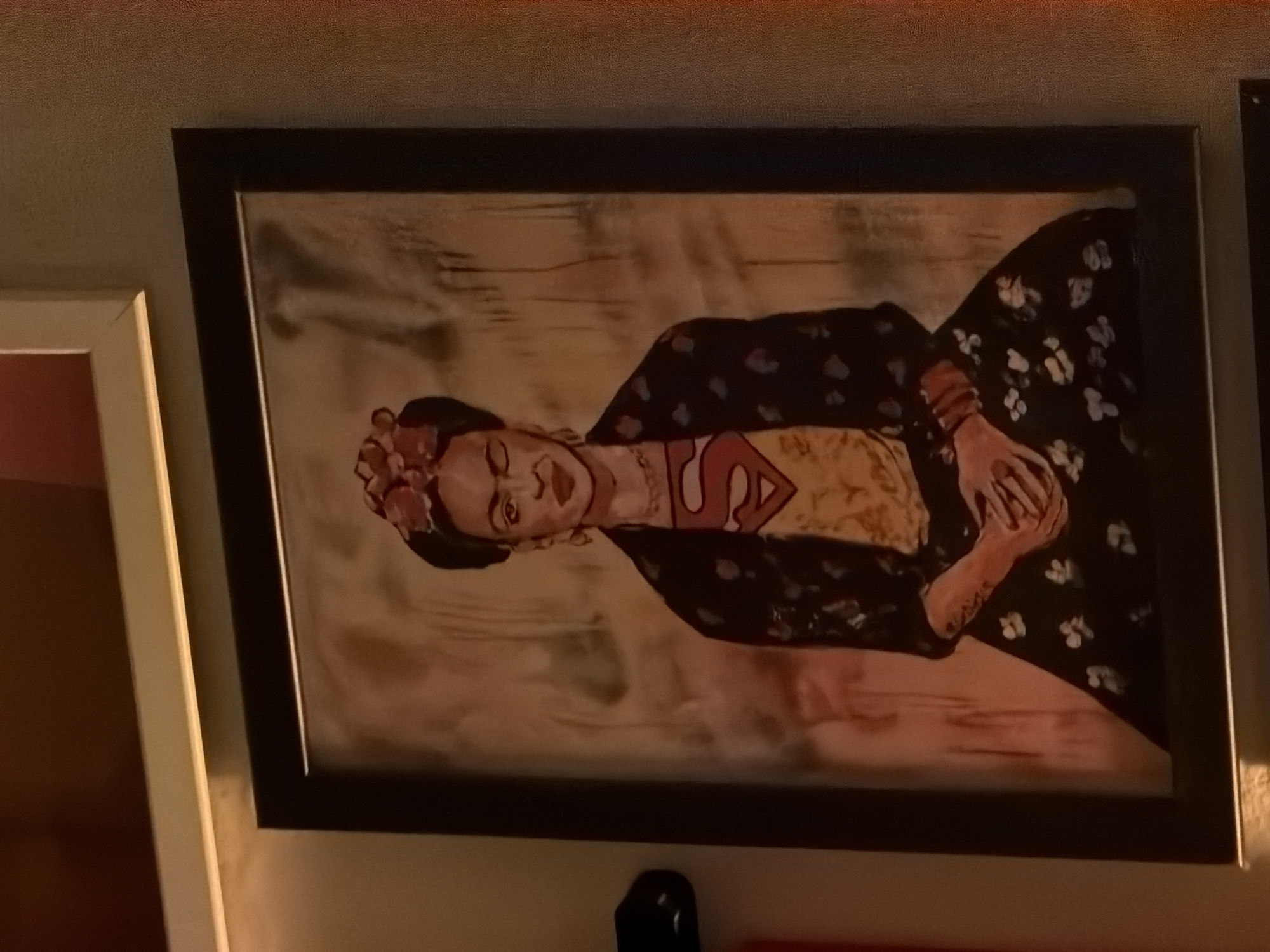 A framed painting is hung on a wall, depicting a figure resembling Frida Kahlo wearing a yellow shirt with a stylized red "S" Superman logo. The figure has flowers in their hair and a dark shawl over their shoulders, resting their hands in their lap. The background is a blurred, indistinct color. A portion of another framed picture is visible to the left of the main painting. There is a black object under the center of the painting. The location in Milan is impossible to determine based on the visual information provided.