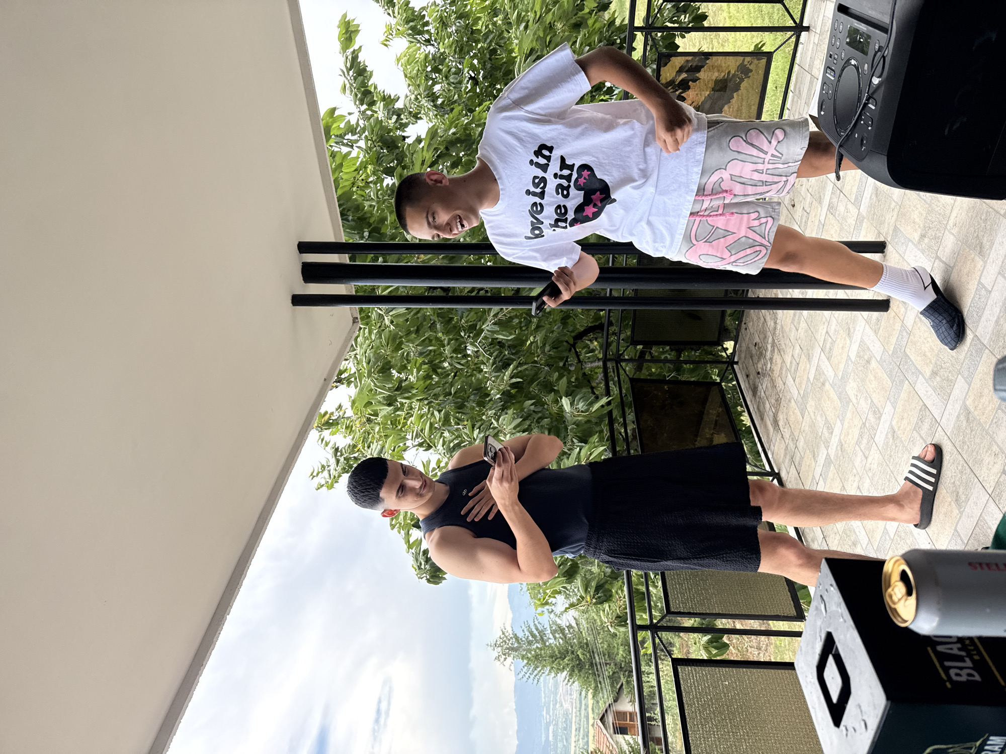 Two young men are standing on a tiled patio area, likely a balcony or rooftop terrace, with a view of a valley and mountains in the background. The man on the left is wearing a dark tank top and a black skirt, while the man on the right is wearing a white t-shirt with the text "love is in the air" and light-colored patterned shorts. Both men are looking at their phones. The man on the right is laughing.
In the foreground, there is a black box with a square opening, a silver can, and a black speaker. The patio is covered by a white overhang supported by black columns. Lush green trees are visible behind the men and the railing of the patio. The sky is cloudy.