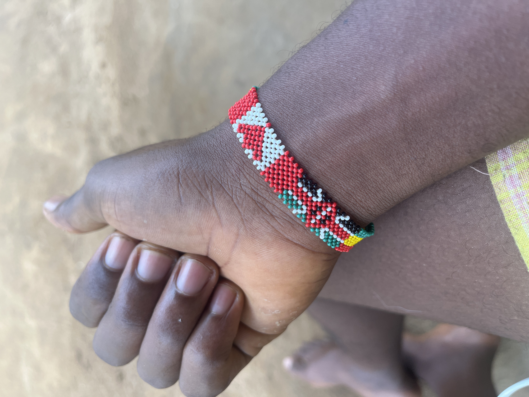 A close-up shows a dark-skinned person's hand, likely a child's, wearing a colorful beaded bracelet. The bracelet's pattern includes red, white, black, green, and yellow beads. The hand is in a partially clenched position. The background appears to be a light-colored sandy surface. Based solely on the visual information, there's no indication of the location being Ikot Ekpene, Nigeria.