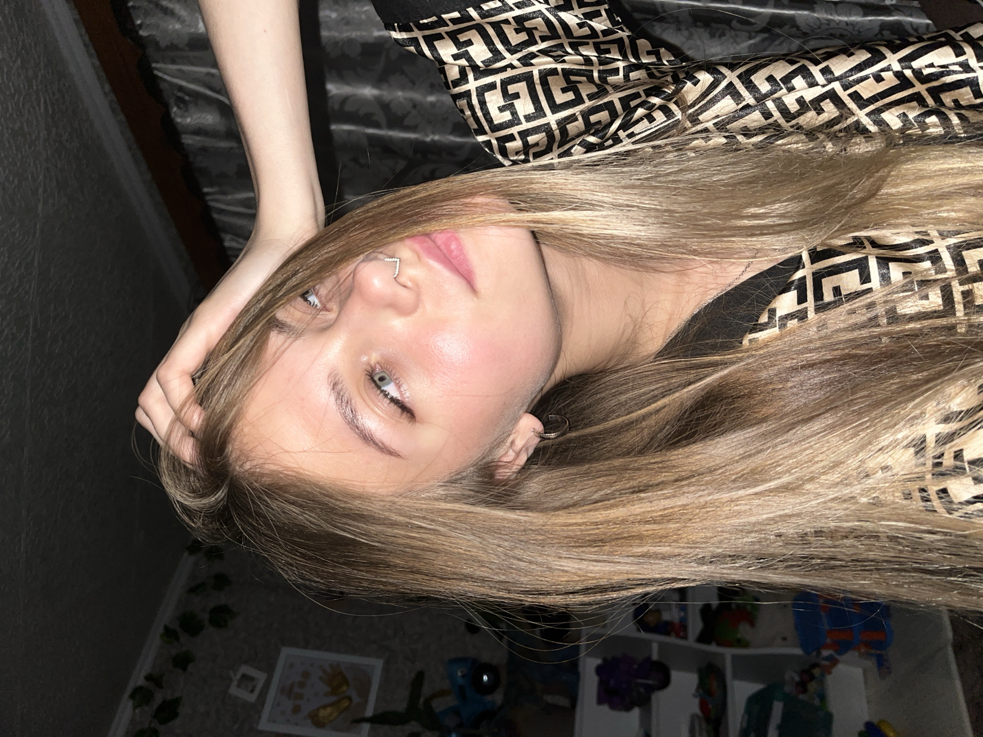 A young female is depicted indoors in a dimly lit personal space, located in the city of Фоки, Russia. She possesses long, straight, light brown hair that partially obscures her left facial profile. Her head is tilted slightly to her left, with her light-colored eyes directed upwards and to her right. A 'V' shaped septum piercing is present, along with a small hoop earring in her visible left ear. Her skin exhibits a sheen, possibly due to illumination or dermal characteristics. Her right hand is raised, with fingers positioned within her hair near the crown of her head. She wears a garment, likely a shirt or robe, characterized by a repeating black and beige geometric pattern. A thin silver necklace chain is visible at her collarbone. The background on the left features a light-colored, possibly textured wall, adorned with an artificial green ivy garland and two framed items, one containing what appears to be golden child's footprint art. Below these, a blurred white shelving unit holds various indistinct objects, including a blue and orange item and a purple floral object. The right background consists of a dark, textured fabric curtain, with a darker structural element visible above it. The scene is illuminated by dim, artificial light.