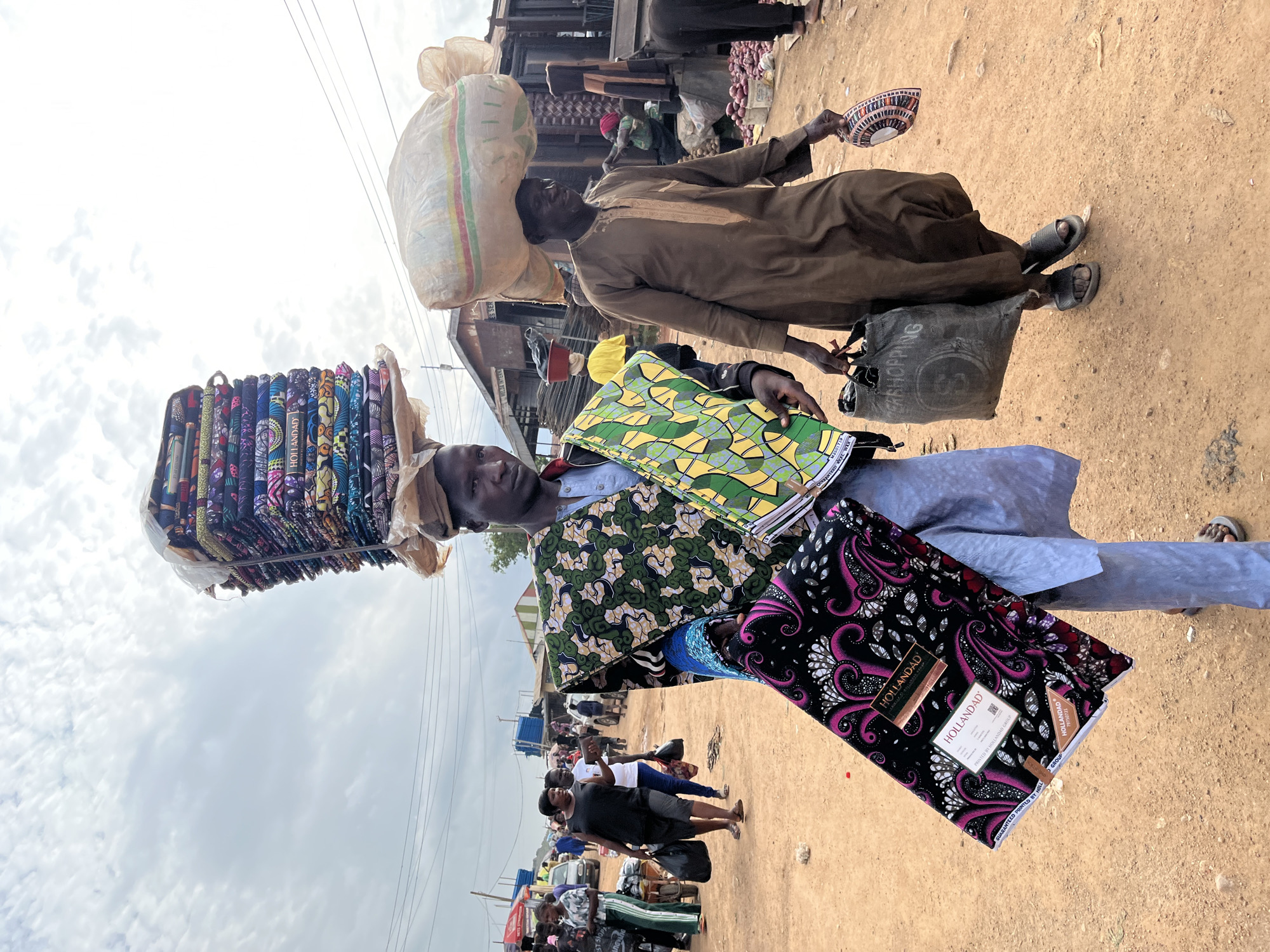 This image depicts a bustling outdoor market scene in Gura Top, Nigeria. The foreground is dominated by two individuals carrying large stacks of vibrant patterned fabrics. The person on the left is balancing an impressive tower of folded textiles on their head, while the person on the right holds several more pieces of fabric, one featuring a striking black background with a pink and white swirling pattern and the "HOLLANDAD" logo clearly visible. To the left of the fabric stacks, another person carries a large, bulging sack of goods on their head, draped in a tan robe.
In the background, more people are visible, engaged in various market activities. Stalls are set up, suggesting a lively commercial environment. The ground is dusty and sandy, typical of an outdoor marketplace. The sky above is partly cloudy, indicating it is likely daytime. The overall atmosphere appears dynamic and energetic, characteristic of a busy market day. Text visible on the fabric packaging includes "HOLLANDAD" and "GROUP", along with some product codes and descriptions like "GUARANTEED PRINTED" and "REAL WAX". The scene captures the daily commerce and the ways goods are transported and displayed in this Nigerian location.