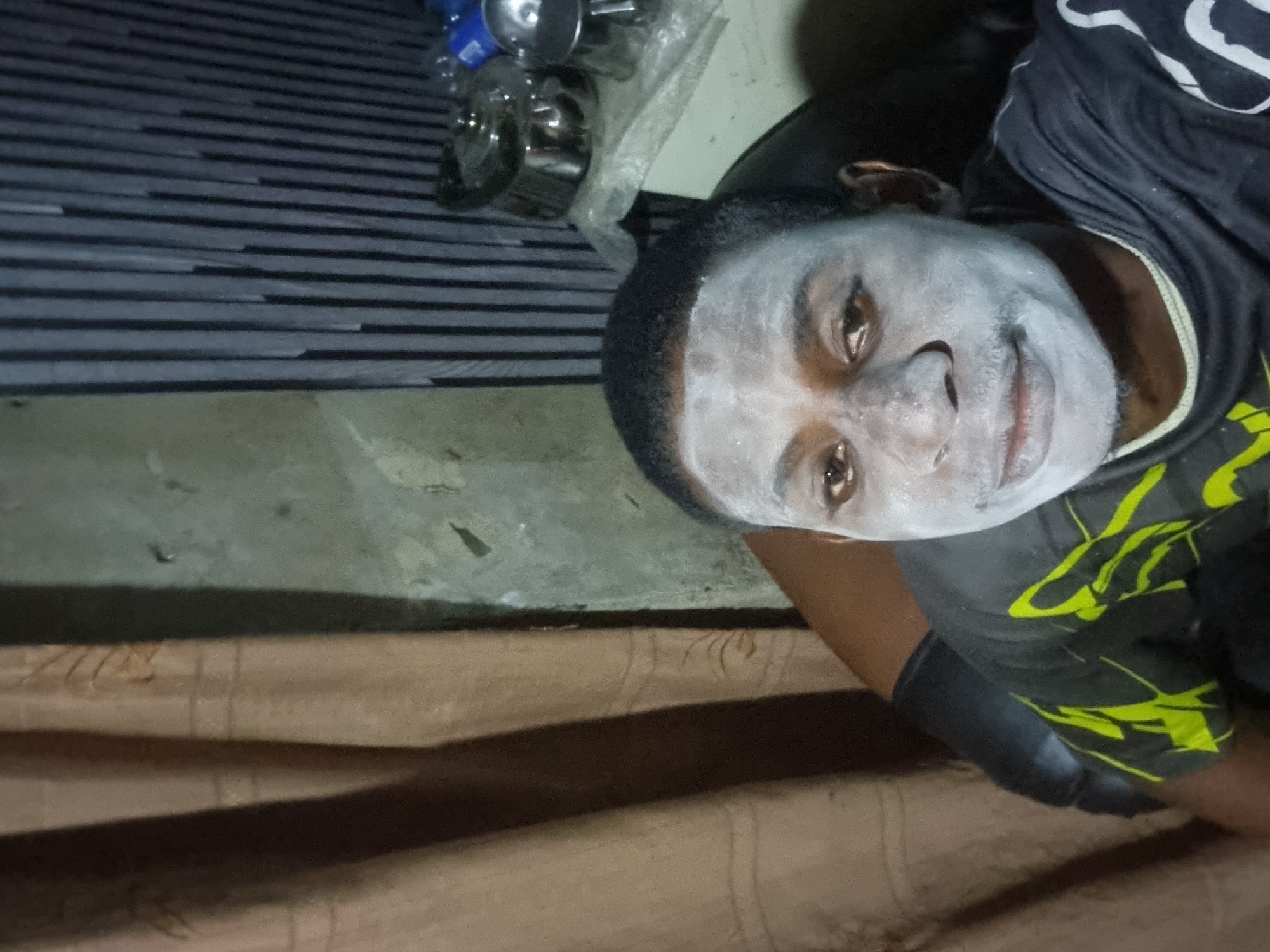 The image, taken in Onitsha, Nigeria, features a single person, likely male, with their face entirely covered in a light-colored, possibly white or pale grey, substance resembling face paint or powder. They have short, dark hair and calm, brown eyes that look directly at the viewer, accompanied by a slight smile. The person is wearing a dark, short-sleeved shirt adorned with neon yellow or green abstract designs on the lower right side.
The setting appears to be indoors, characterized by dim lighting, suggesting either evening or a space with limited natural light. In the upper left background, various objects are visible, including several metallic items that resemble kitchenware (pots or bowls), some blue plastic bottles, and an item partially wrapped in clear plastic. Below these objects is a dark, striped surface, possibly a patterned floor mat or tiles. Adjacent to this and below the metallic objects, the floor transitions to a plain, greyish, concrete-like surface. Below the person, a textured brown surface, possibly fabric or a cushion, can be seen.
The image is rotated 90 degrees clockwise, giving the impression that the person is positioned horizontally or the photo was taken with the camera on its side. The activity shows the individual posing for the camera, highlighting the facial application, which could be for cultural practices, performance, or personal expression. The person's expression conveys a sense of ease and a subtle hint of amusement. There is no visible text in the image.