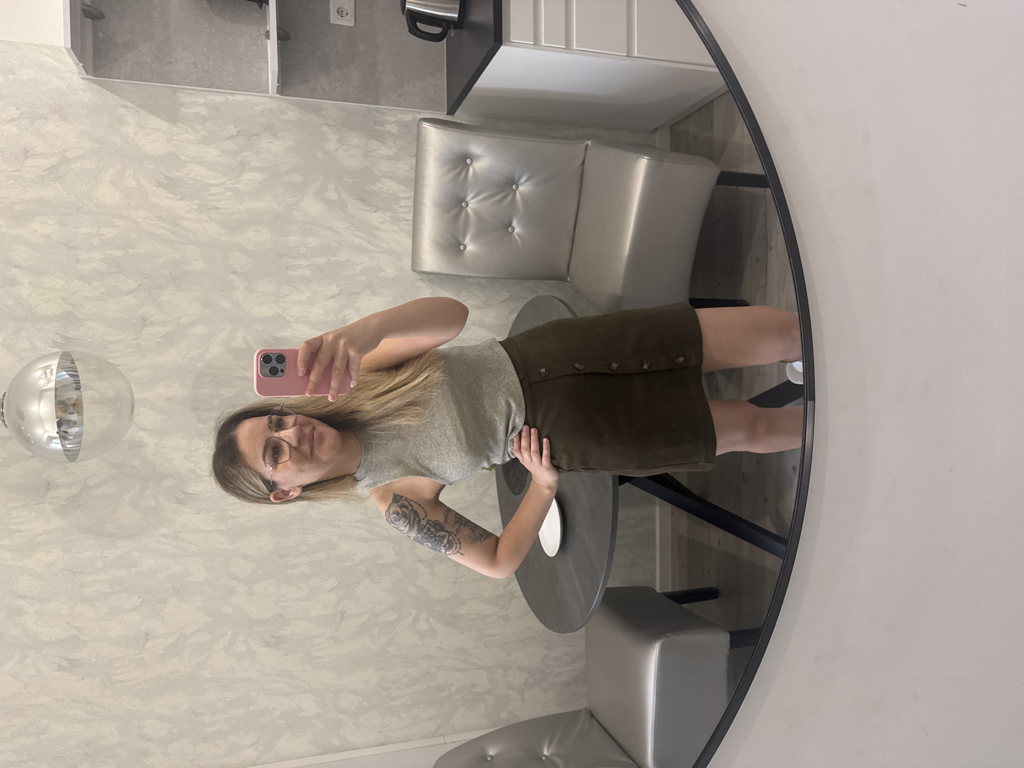 A woman is taking a selfie in a mirror. She has blonde hair and is wearing glasses, a grey tank top, and a dark green skirt. She has a tattoo on her left arm. She is standing in what appears to be a dining or kitchen area with a round table and two chairs. There is also a white cabinet with a kettle on top, a shelving unit, and a wall outlet visible. The room has textured grey and white walls. The reflection in the mirror shows a silver spherical light fixture hanging from the ceiling. The image does not provide any information to determine its location in Budapest, Hungary.