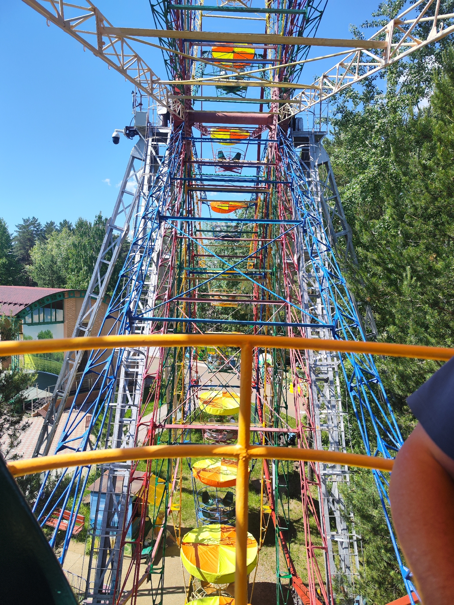 The image shows a view from a Ferris wheel at an amusement park. The Ferris wheel is brightly colored with red, blue, yellow, and green metal struts. The gondolas are yellow and orange. The Ferris wheel is located in a wooded area with a clear blue sky above. In the background, there is a building with a red roof and green windows, and some trees. The image is taken from the perspective of someone on the Ferris wheel, looking down and into the structure. A person's arm is visible in the foreground on the right side of the image.