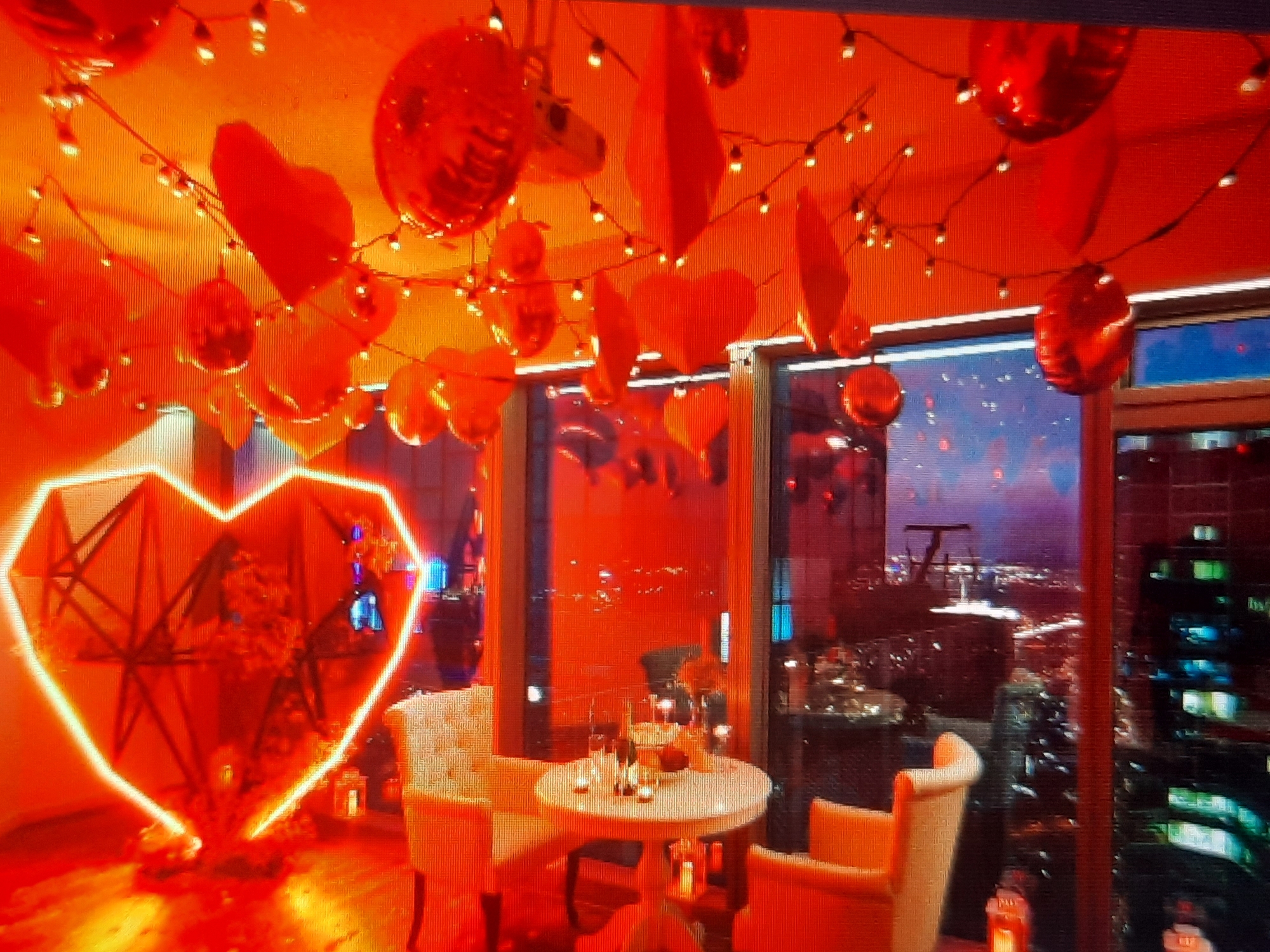 The image is an indoor scene, likely a shop window display, featuring a vibrant orange and red color scheme. Prominently displayed in the upper portion of the image is a large, illuminated heart shape outlined in bright white or yellow light, with a dark, skeletal structure visible within its borders. To the right, a string of small, white lights is intertwined with numerous red, reflective, bulbous ornaments, some appearing to be shaped like hearts or mushrooms.
In the lower half of the image, there are layered shelves or displays. On the left, a white, multi-faceted object, possibly a display stand or part of a fixture, is visible. The shelves themselves appear to contain various objects and reflections, including hints of blue and purple light, suggesting other displays or lights in the background. The overall impression is one of a festive or decorative setting, possibly related to a holiday or special event.
The image does not provide any information about the city of Rosenheim, Germany, nor does it depict any people or specific actions or interactions. The description is based solely on the visual elements present in the photograph.