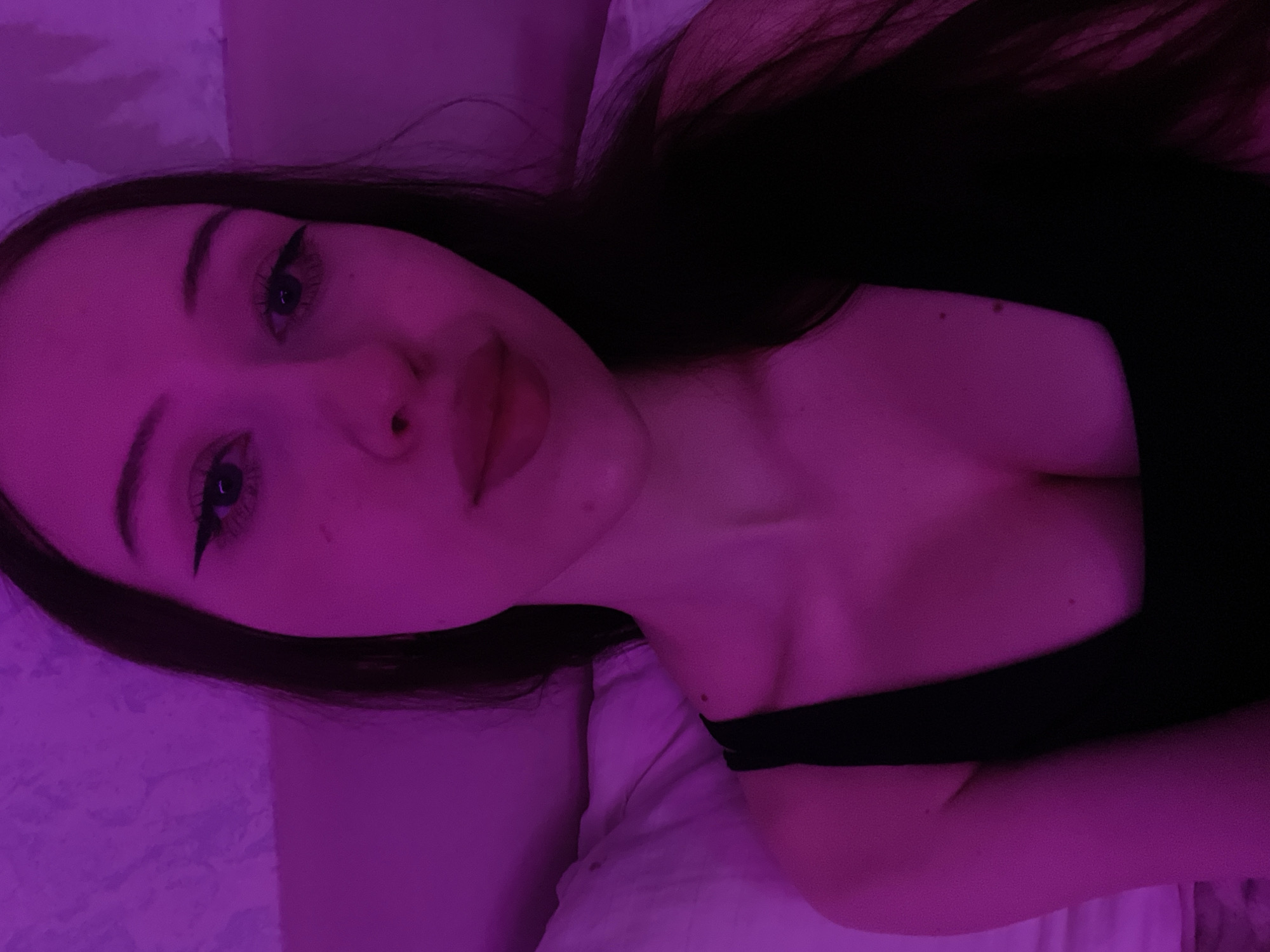 The image shows a close-up, selfie-style shot of a young woman with long dark hair. She is wearing a black tank top. The lighting is predominantly pink, suggesting artificial light from a neon lamp or colored bulb. The background appears to be a textured wall and a white pillow. The woman's face is angled towards the camera, with her eyes looking directly at the viewer. She has winged eyeliner and full lips. There is no indication of the city of Votkinsk, Russia, in the image.