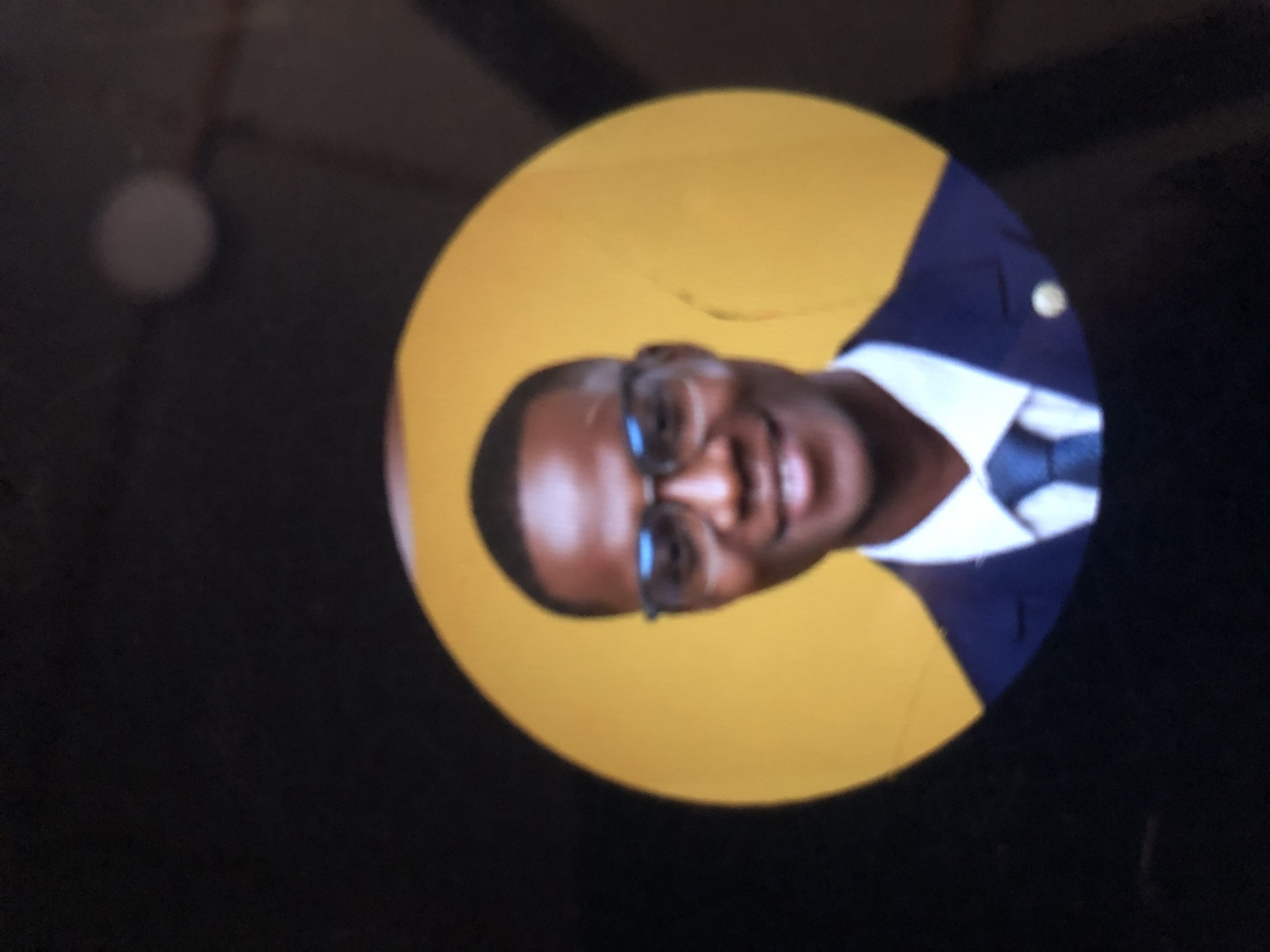 The image shows a man with dark skin, wearing glasses, a white shirt, a blue tie, and a navy blue blazer with a gold pin on the lapel. He is smiling and his face is facing the viewer, but rotated at an angle. The background is a yellow circle, and the rest of the image is a dark black or gray. The location is Unknown, Unknown.