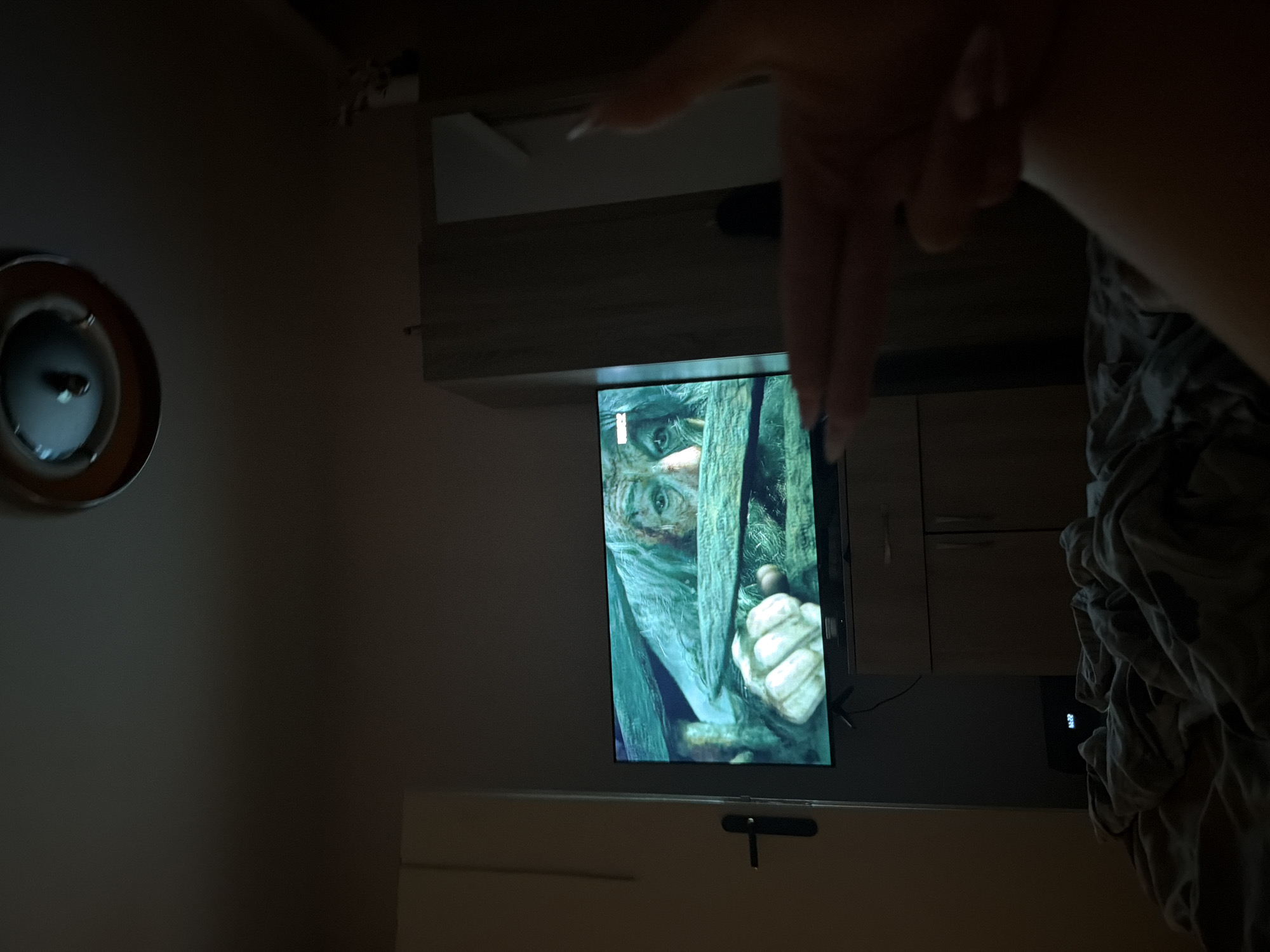 The image depicts a dimly lit bedroom interior. A large flat-screen television is positioned on a cabinet, displaying a close-up of an elderly person's face, appearing to be from a film or show. The person on the screen has pale skin, white hair, and intense eyes, peering through what appears to be wooden bars. A hand, with painted nails, is held up in the foreground, forming a gun gesture towards the television. The bed is covered with rumpled grey bedding. A wardrobe is visible in the background to the right of the television. The scene is consistent with a domestic setting, likely a bedroom, with no specific identifying features of Stara Zagora, Bulgaria.