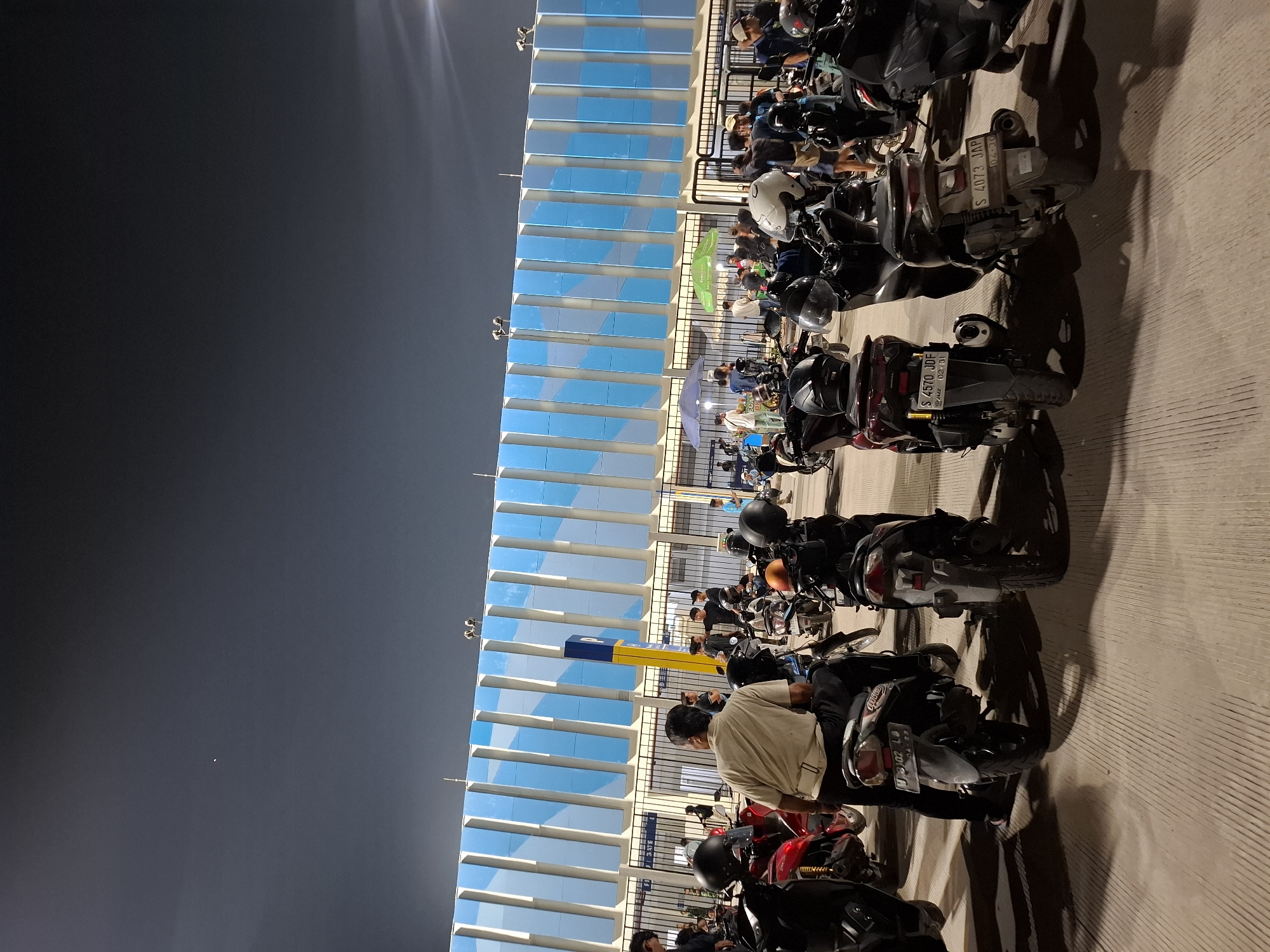 An exterior, nighttime, eye-level medium shot captures a wide paved area in front of a modern building with blue and white vertical panels. Numerous motorcycles are parked, with several people standing or sitting on them. In the foreground, a man in a light-colored shirt and dark pants sits on a motorcycle. To his right, another motorcycle and rider are visible. The building's facade features a repeating pattern of blue and white rectangular panels, with a tall yellow pole displaying a blue "P" sign. The sky is dark with a single bright light visible. Several food stalls with umbrellas are set up in the middle ground. The scene suggests a gathering of people and vehicles, possibly at an event or a popular nighttime location in the city of Perumda Deket, Indonesia.