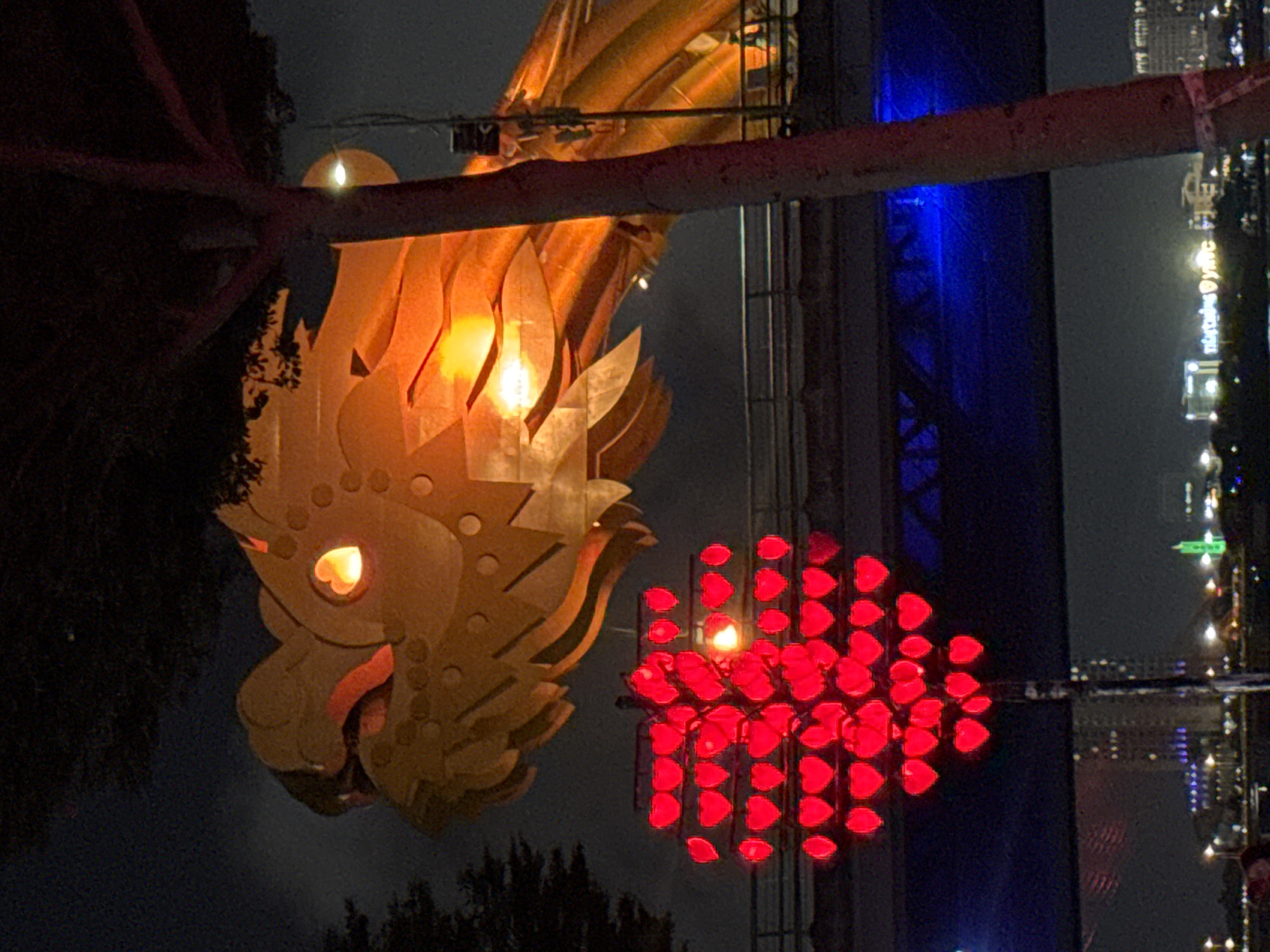 The image captures a nighttime scene in Đà Nẵng, Vietnam, featuring illuminated decorations near the Dragon Bridge. A golden dragon head with a glowing eye is on the left, and a red heart-shaped light display is to its right. In the background, there's a building illuminated with blue light and distant city lights across the river, reflecting on the water. Trees frame the bottom of the image.