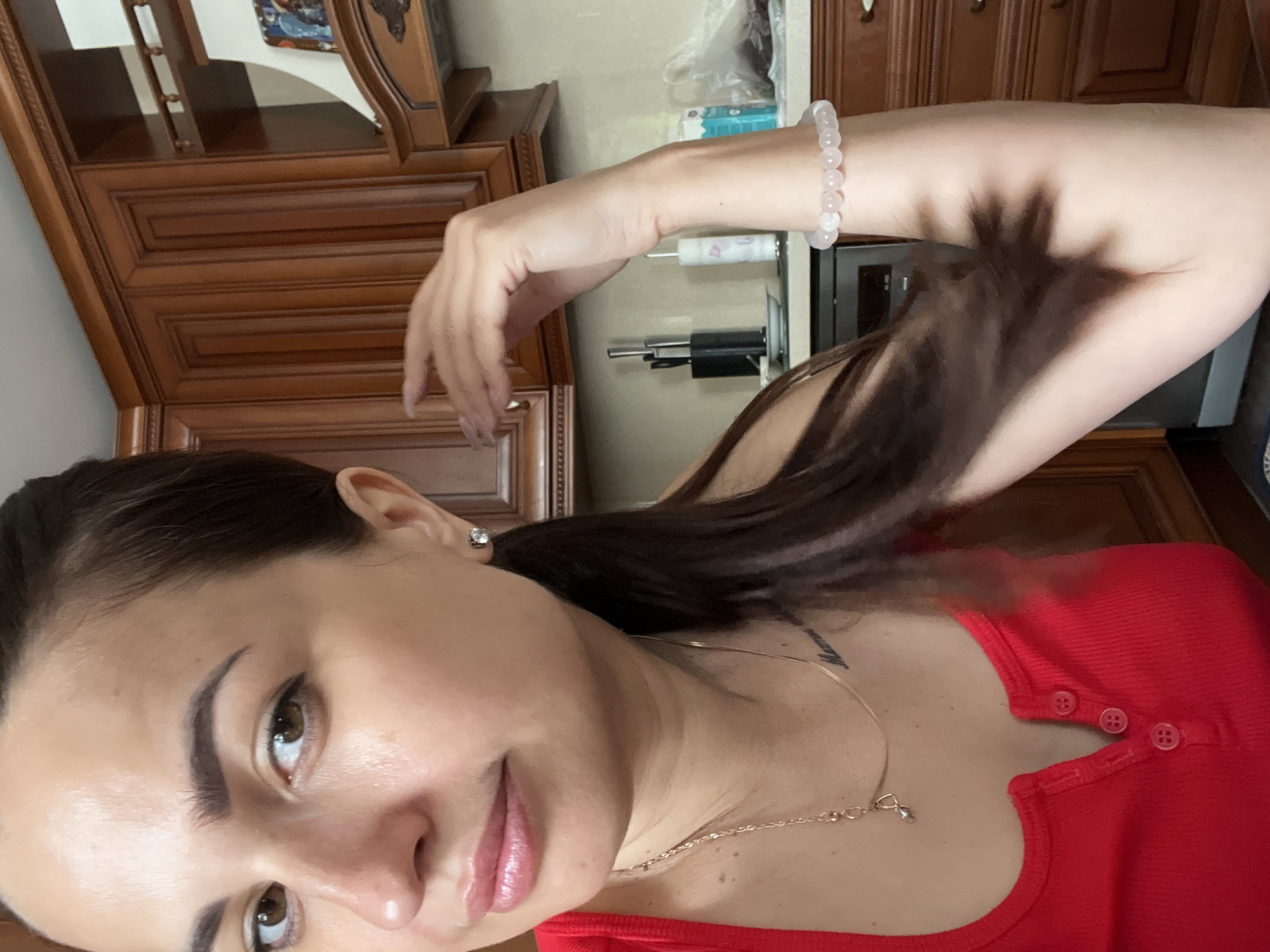 An individual, identified as female by facial features and attire, is captured in a close-up, waist-up portrait. She has dark brown hair, styled in a ponytail, with visible strands framing her face. Her skin appears light-toned with a slight sheen. She is wearing a vibrant red top with a buttoned neckline. A delicate gold necklace with a small pendant adorns her neck, and a tattoo is visible on her upper chest area. Her right arm is bent, with her hand near her head, and she is wearing a bracelet composed of light pink beads on her wrist. The background suggests an indoor setting, likely a kitchen, with warm-toned wooden cabinets visible. The lighting is moderate, casting some shadows. The image is taken from a slightly upward angle.