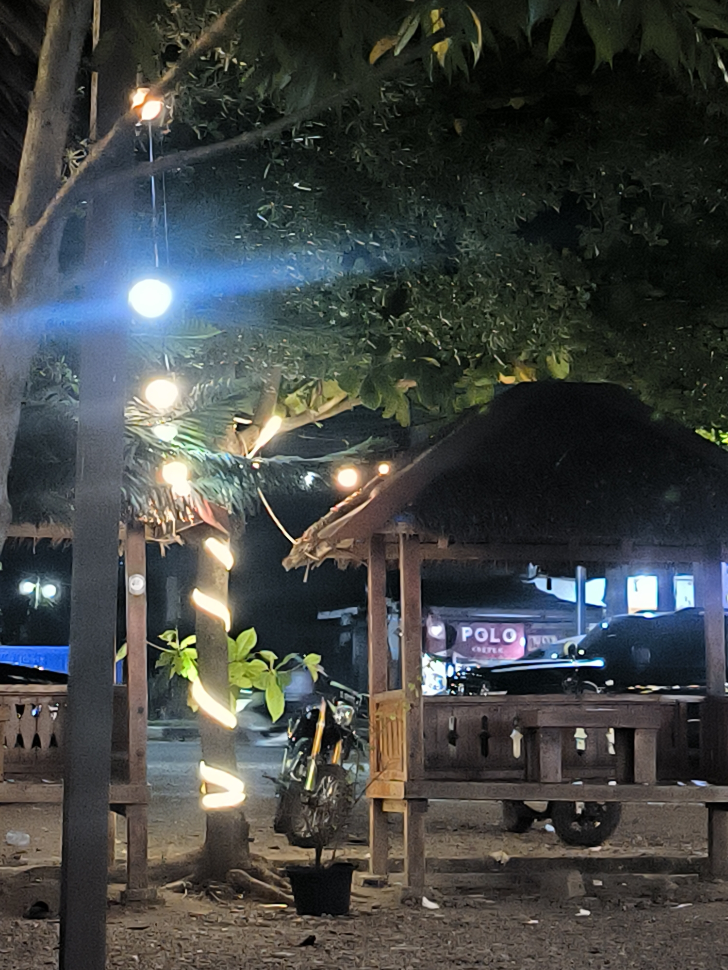 The image, taken at night, depicts a scene in Titibaro, Indonesia. A small wooden gazebo with a thatched roof occupies the right side of the frame, partially obscuring a storefront with the word "POLO" visible on a sign. A motorcycle is parked to the left of the gazebo, near a tree that is wrapped in decorative string lights. Overhead, string lights are suspended among the tree branches. A blue tarp is visible in the background. The ground is covered in gravel.