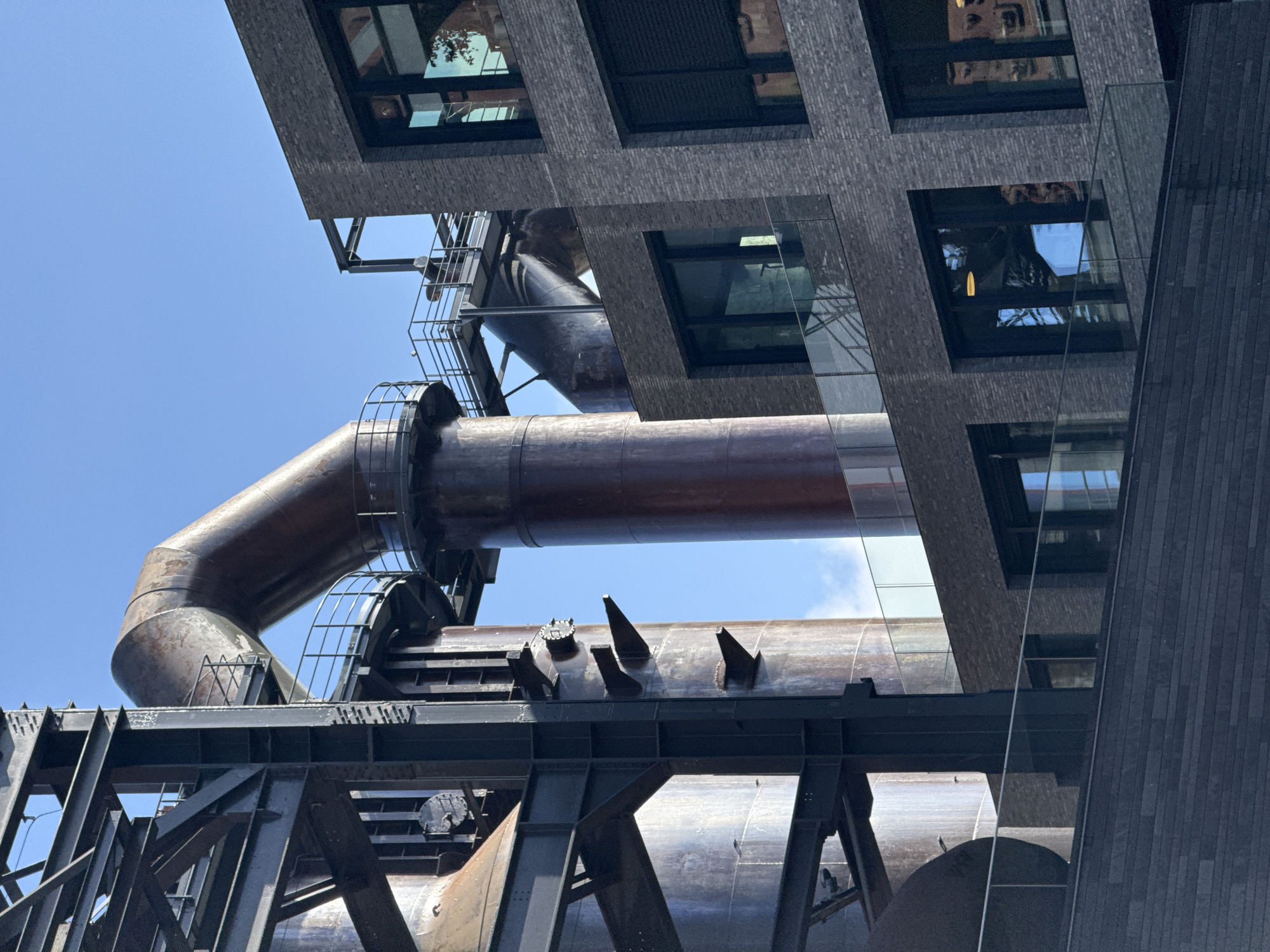 This image depicts a juxtaposition of preserved industrial architecture and modern urban development, specifically located in the Belval district of Esch-sur-Alzette, Luxembourg.
The scene is dominated by two large, weathered steel structures characteristic of decommissioned blast furnaces. On the left, a tall, cylindrical section of a furnace, heavily patinated with rust and dark streaks, is supported by a robust framework of dark-painted or oxidized steel girders and beams. Several conical protrusions and circular access points are visible on its surface. In the center-background, another substantial rust-colored cylindrical structure rises, topped by a large, arching, rusty metal pipe or duct that extends towards the right. Multiple metal platforms with black railings provide access points around these industrial elements.
To the right, a modern multi-story building is visible, featuring a dark grey brick facade laid in a regular pattern. It presents large, rectangular windows with dark frames, some reflecting the blue sky and surrounding elements. Several levels incorporate balconies or terraces, distinguished by clear glass railings. On an upper floor, one window is equipped with a dark, possibly retracted, exterior roller blind. A lower section of the modern construction exhibits a dark, horizontally paneled cladding.
The sky is clear blue with minimal, faint white cloud wisps in the upper-right. No individuals or active processes are discernible within the frame. The lighting suggests a bright, sunny daytime environment.
