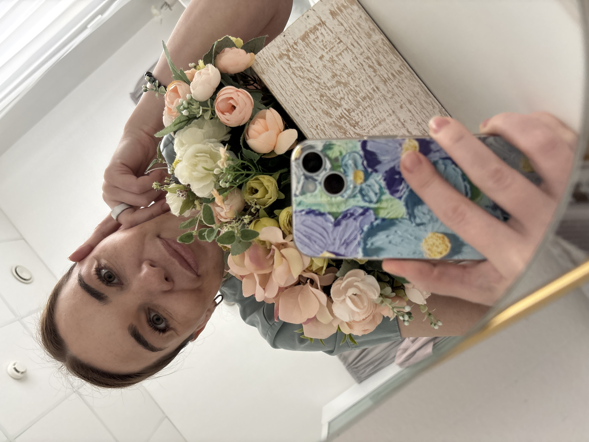 A selfie is being taken in a mirror. A woman is holding a floral arrangement and a smartphone with a blue floral case. The phone is positioned to capture the reflection in the mirror. The environment appears to be indoors, likely in a room with a light color scheme, and possibly in Romanovka, Russia.